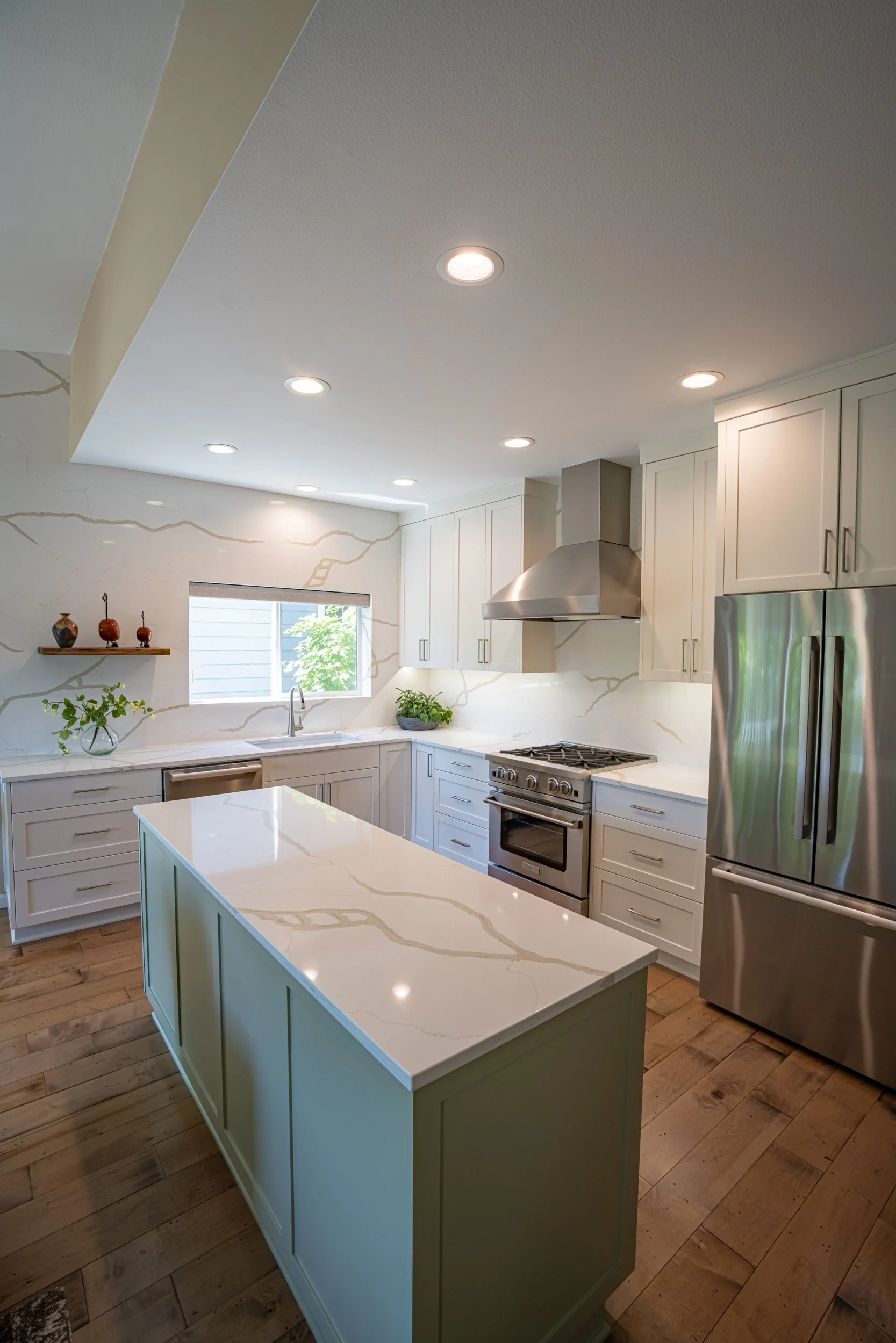 Modern kitchen with white cabinets, marble countertops, stainless steel appliances, and wooden floors, illuminated by ceiling lights and natural light from a window.