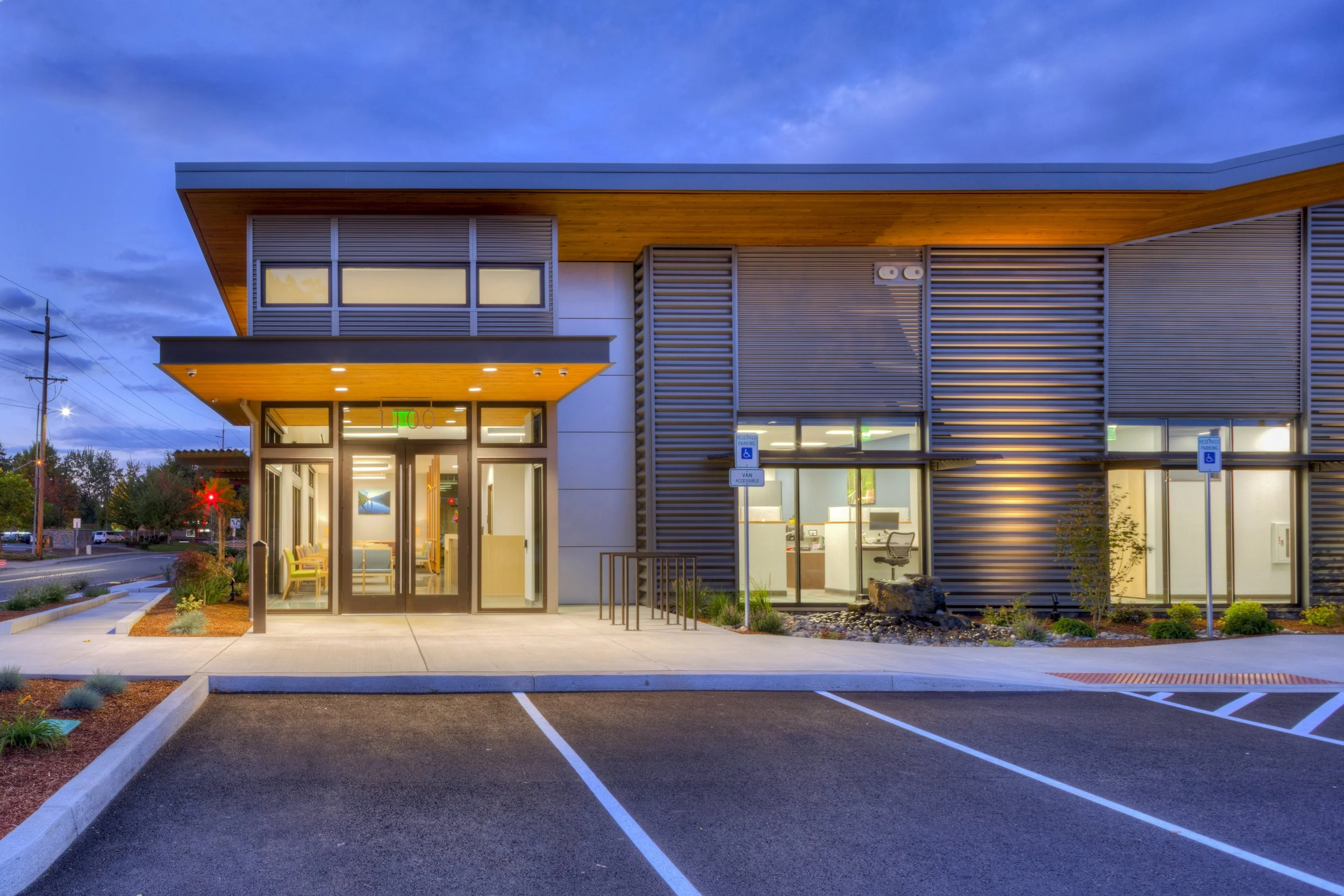 A modern commercial building with illuminated windows and a parking lot in the foreground during dusk. The building features horizontal metal siding and wooden accents with an overhanging roof.