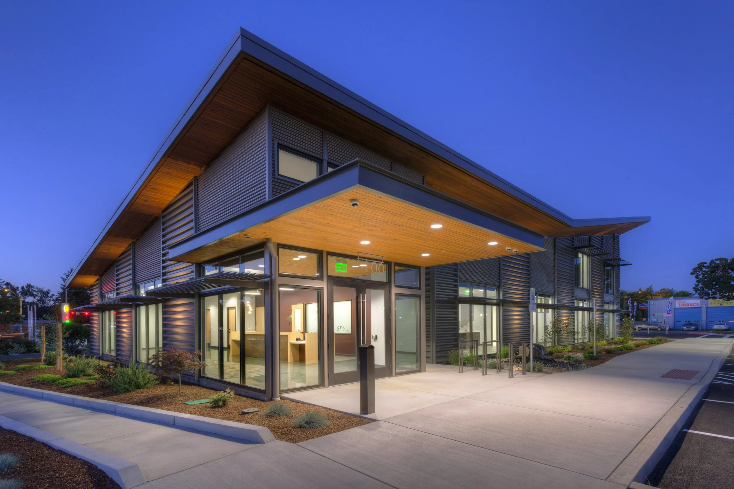A modern commercial building with large glass windows and a wooden overhang, illuminated at dusk, with landscaped surroundings and parking lot.