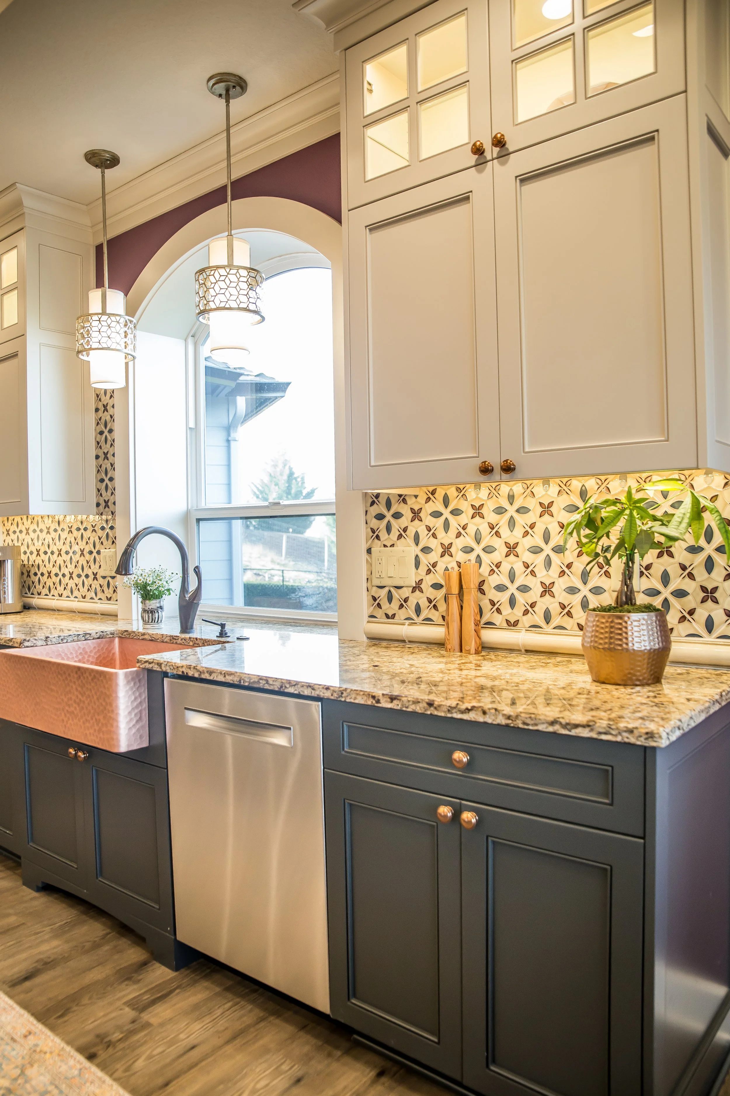A kitchen with dark blue lower cabinets, a granite countertop, and white upper cabinets with glass-paneled doors. There are two pendant lights hanging from the ceiling, a window with a view outside, and a copper-colored farmhouse sink. A potted plant and a small plant are on the countertop.
