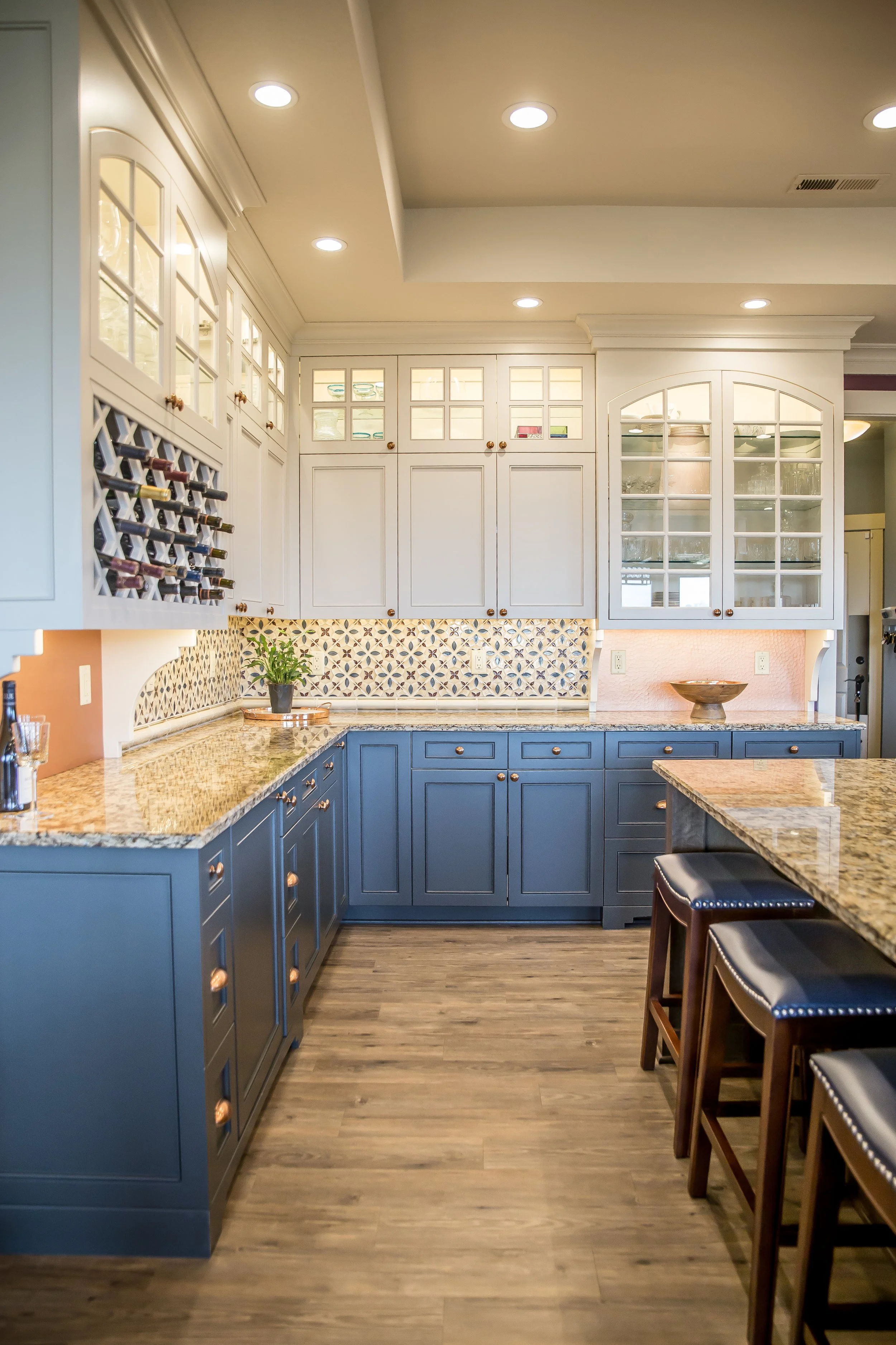 A kitchen with blue lower cabinets, white upper cabinets with glass doors, a granite countertop, a backsplash with a geometric pattern, bar stools with Black seats and wood legs, and recessed ceiling lights.