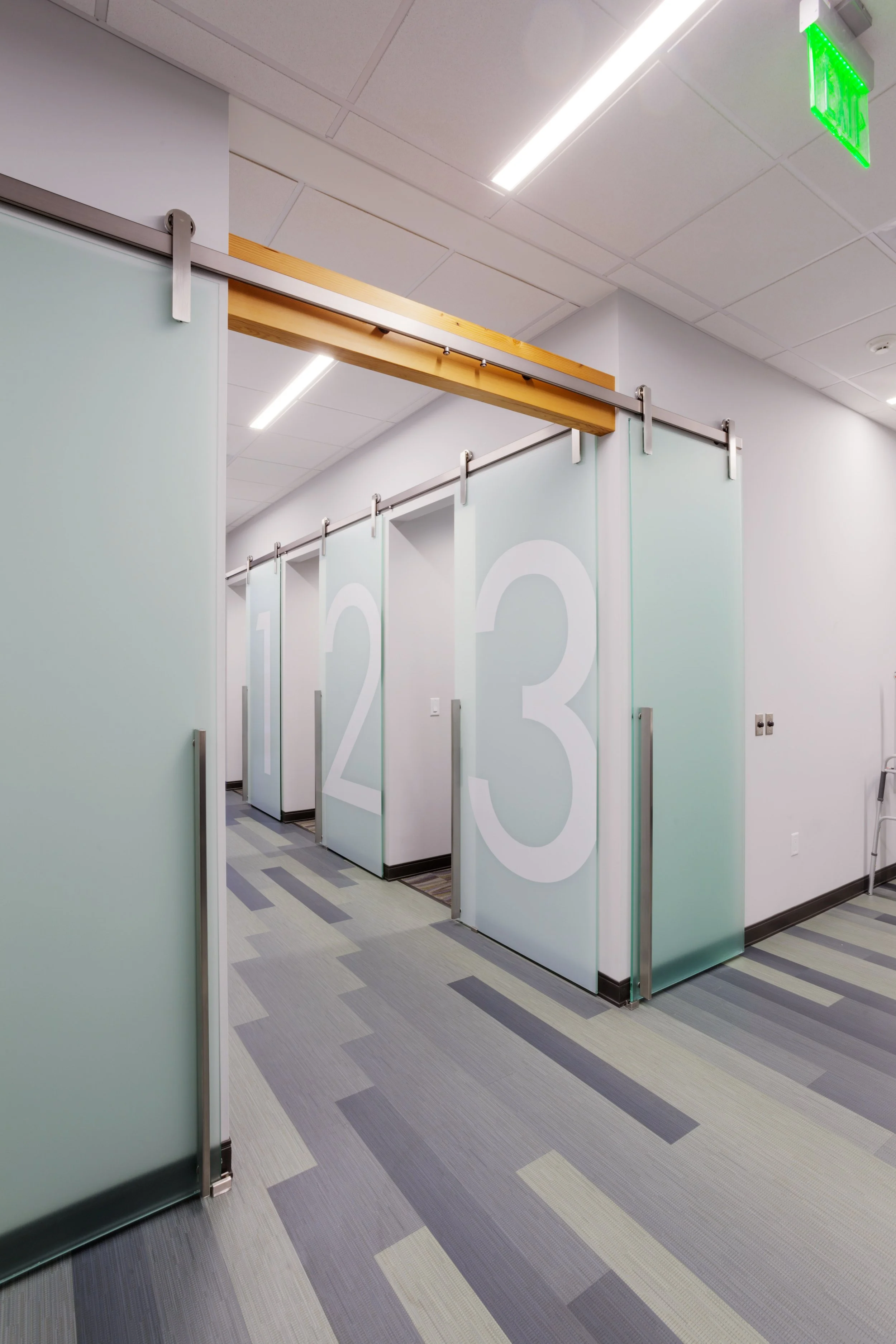 A hallway with green frosted glass partitions numbered 1, 2, and 3, and a polished wood sliding door track on a ceiling with bright lighting and modern gray, purple, and beige patterned flooring.