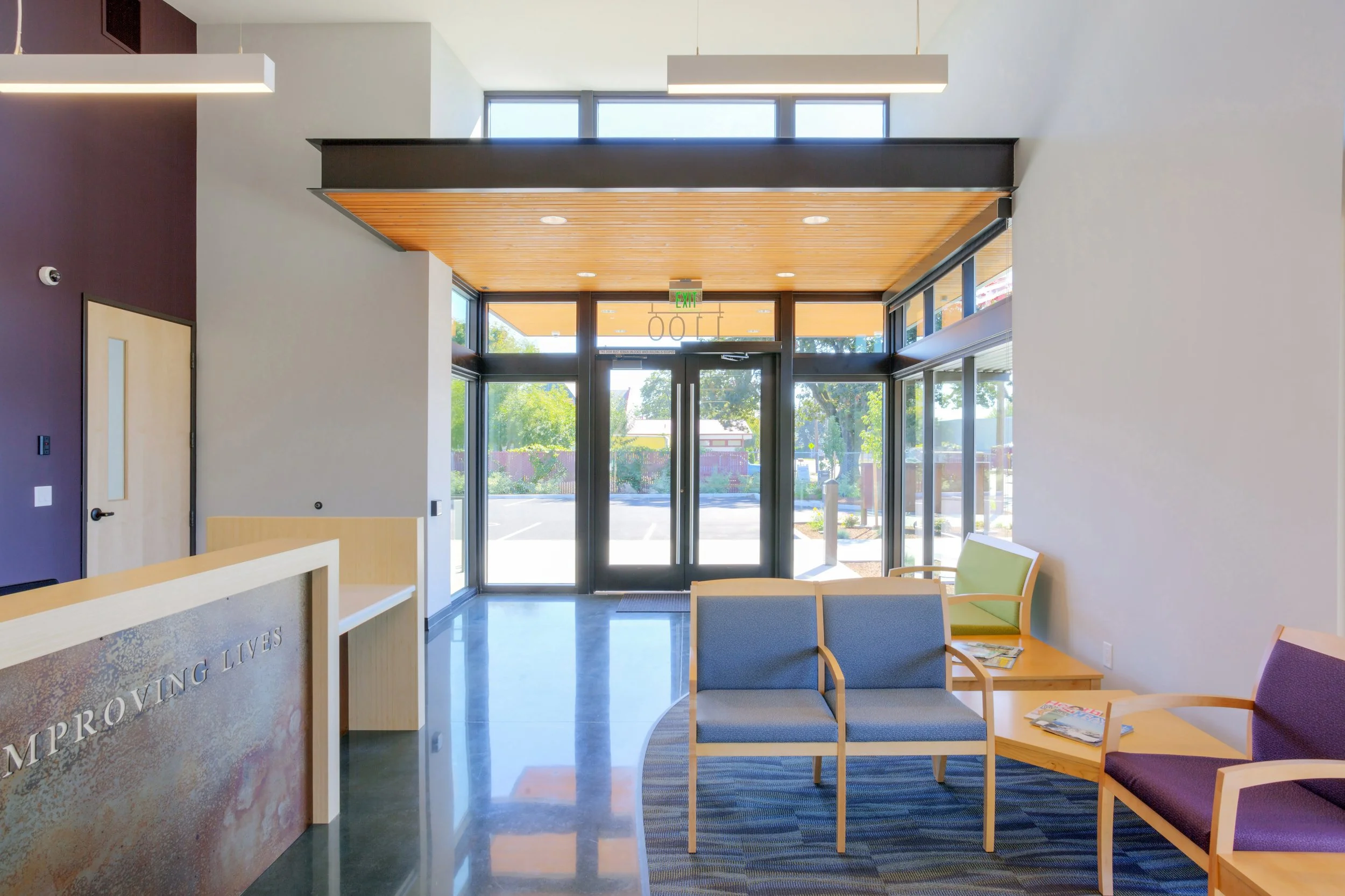 Interior view of a modern building lobby with glass entrance doors, seating area with chairs and tables, and natural light from large windows.