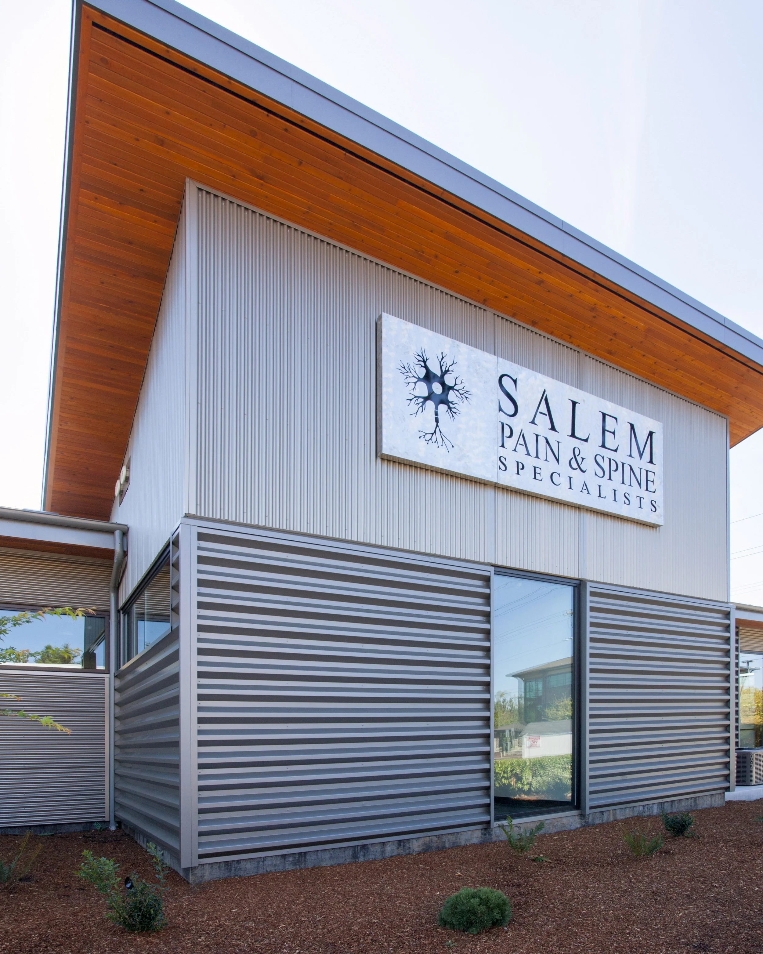 Close-up view of a modern commercial building with a metal exterior, wooden soffits, large glass windows, and a sign for Salem Pain & Spine Specialists, surrounded by landscaped ground with small plants and mulch.