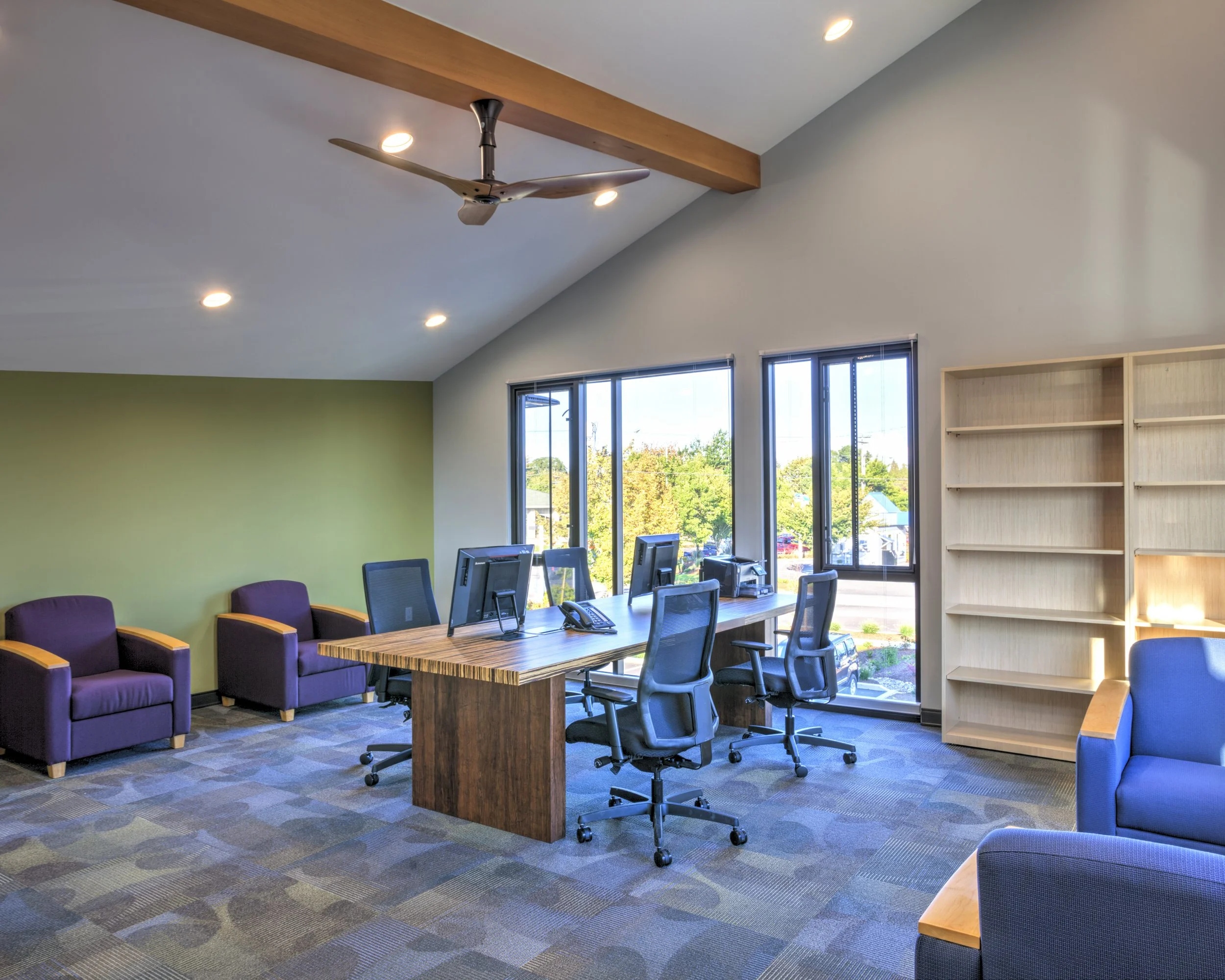 Modern conference room with large windows, a wooden table with multiple office chairs, purple armchairs against the wall, a bookshelf, and a ceiling fan, with sunlight streaming through the windows.
