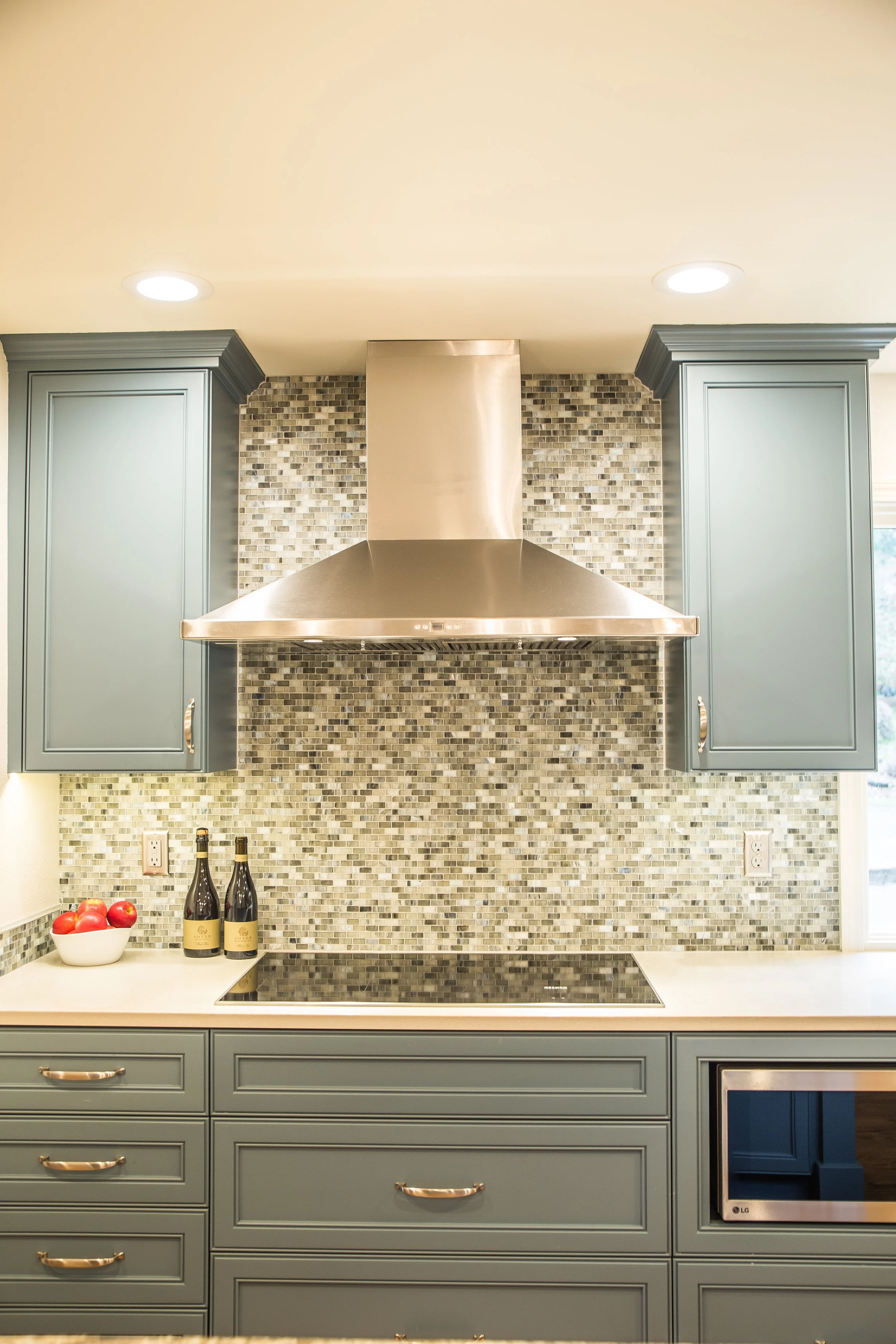 Kitchen with blue cabinets, a stainless steel range hood, a mosaic tile backsplash, a black electric cooktop, a white countertop, a bowl of red apples, and two bottles of wine on the counter.