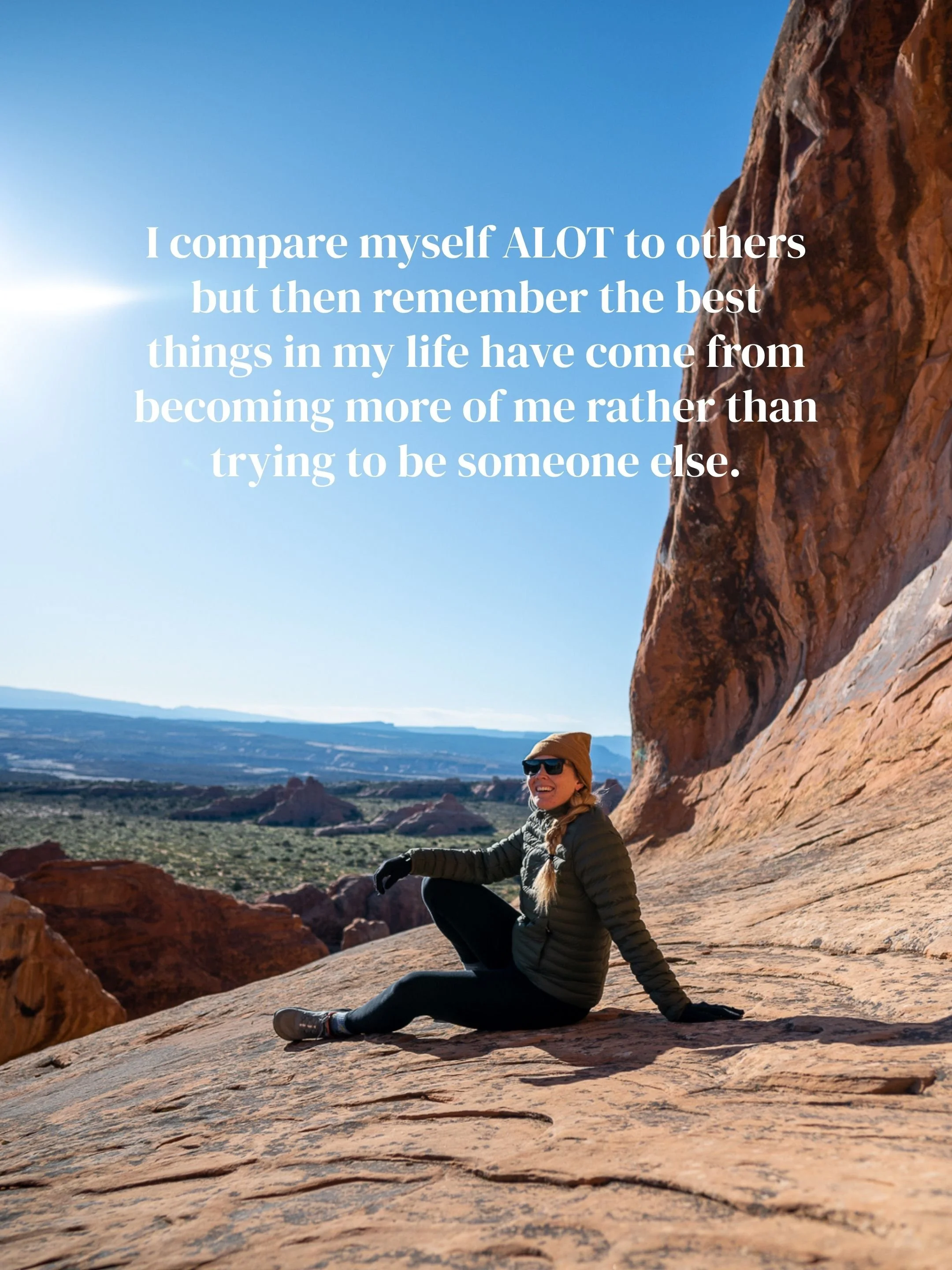 A woman sitting on a large rock formation in a desert landscape, wearing sunglasses, a beanie, a puffer jacket, and hiking shoes, with a clear blue sky and distant red rock formations in the background.