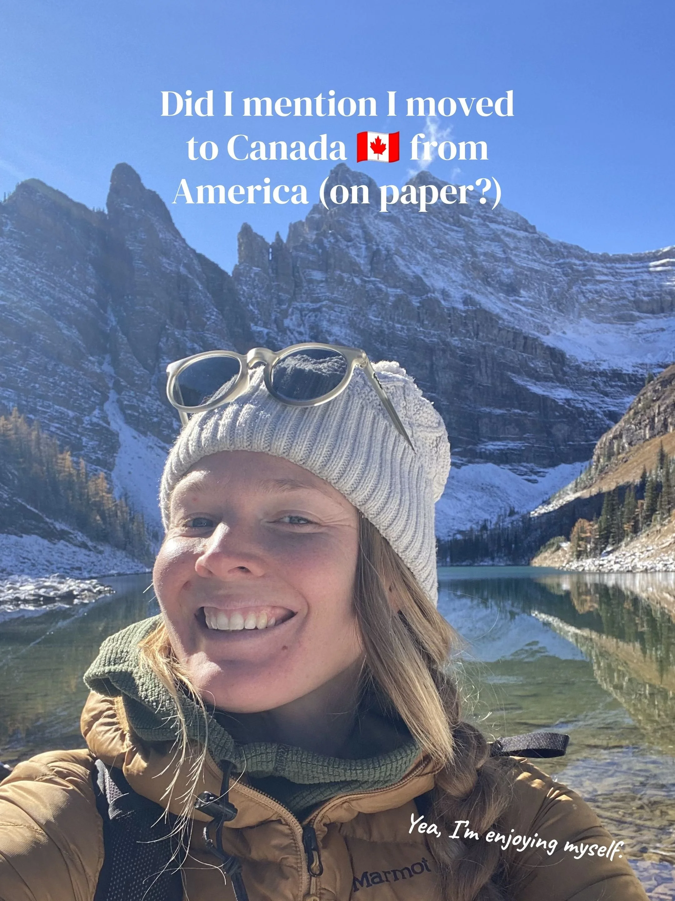 A woman smiling outdoors with snow-covered mountains and a body of water in the background, wearing a beige jacket, a cream-colored beanie, and sunglasses resting on her head. Text overlay says, 'Did I mention I moved to Canada 🇨🇦 from America (on 