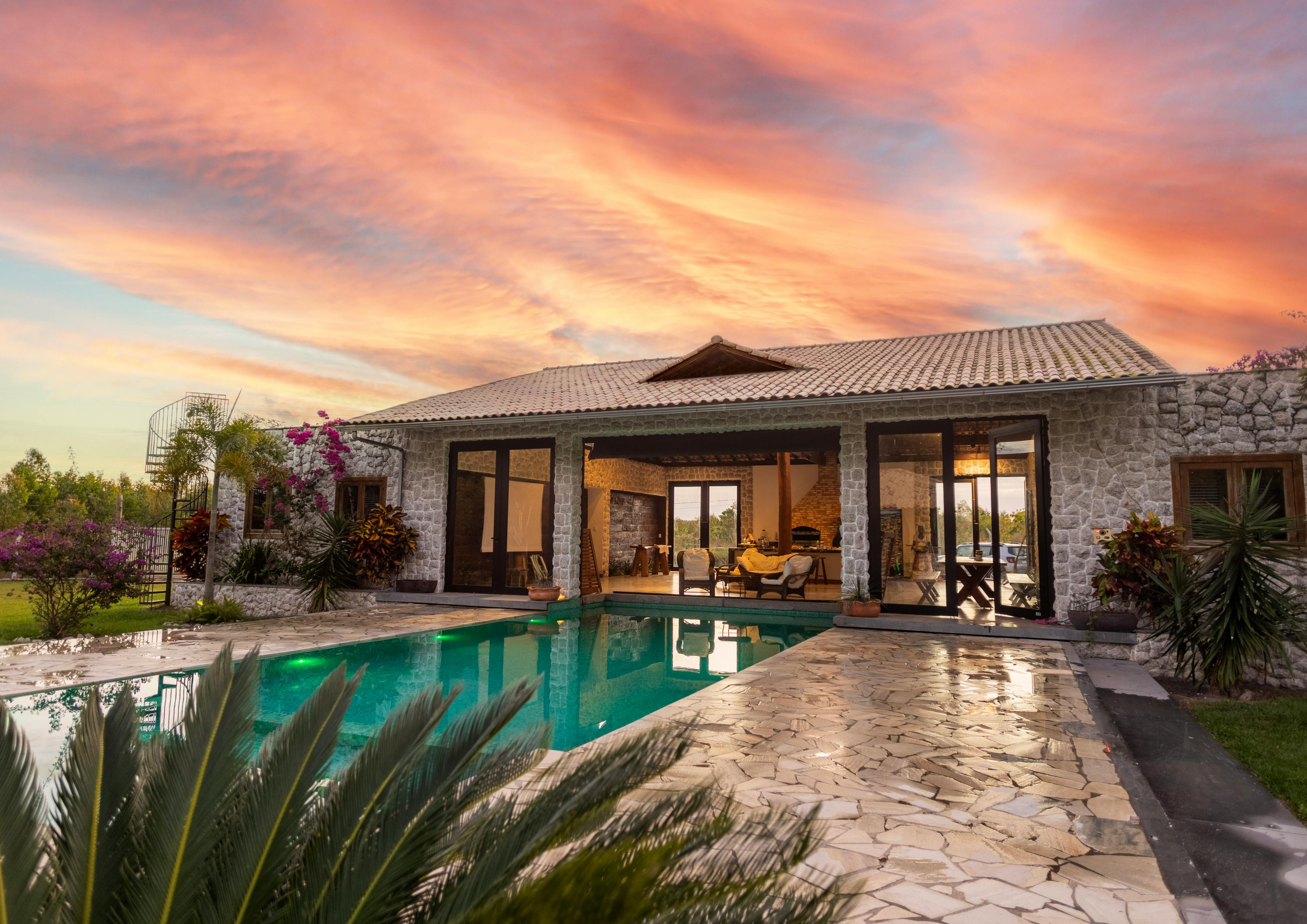 A house with a stone exterior, large glass doors, and a tiled roof, surrounded by plants and flowers, with a swimming pool in the foreground and a colorful sunset sky above.