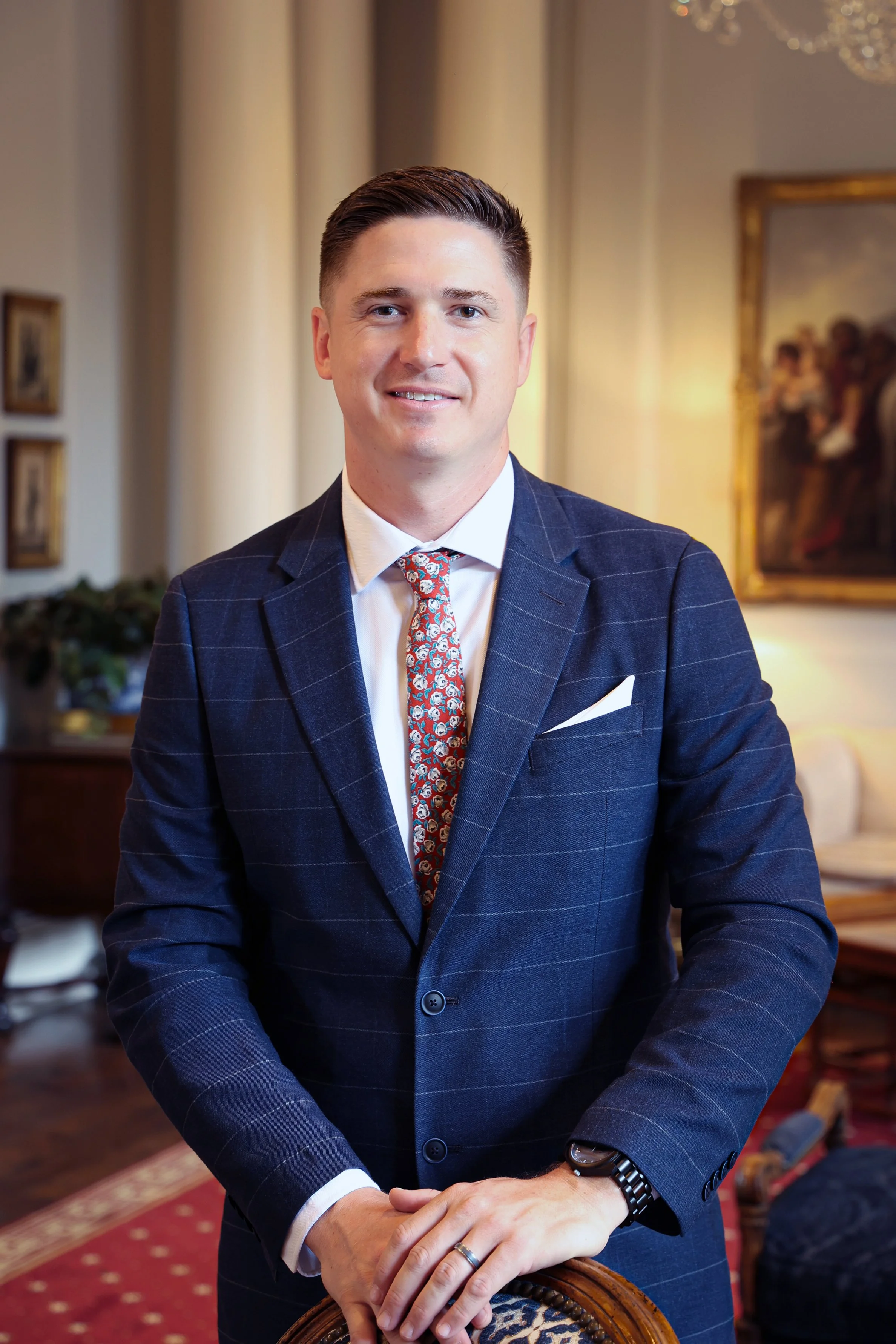 A well-dressed man with short dark hair, wearing a navy blue suit with a windowpane pattern, a white shirt, and a colorful patterned tie. He is standing indoors, with a background that includes framed artwork and warm lighting.