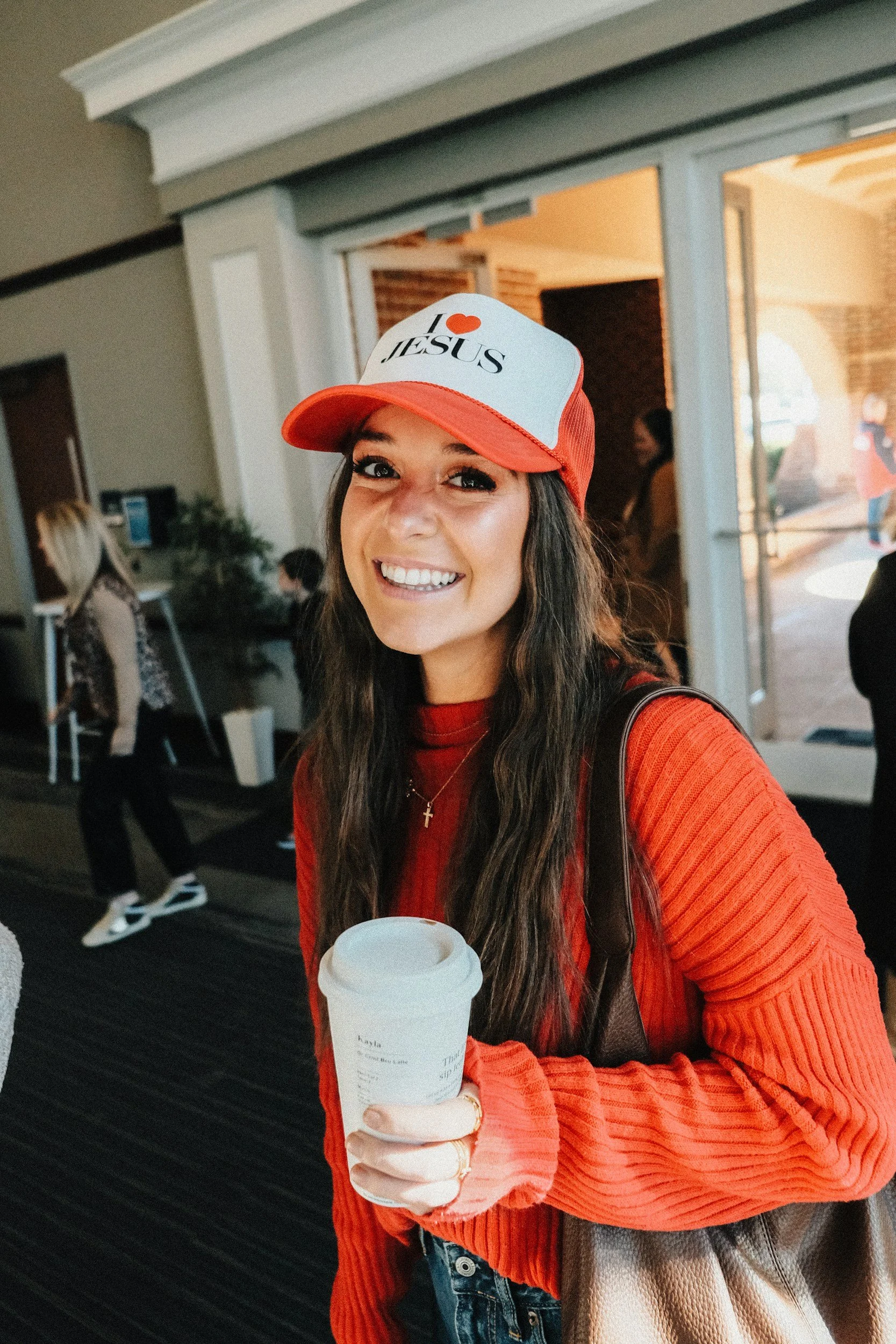 Young woman smiling and holding a coffee cup at an indoor or outdoor location, wearing a red-orange sweater, a baseball cap that says "I love Jesus," and a cross necklace.