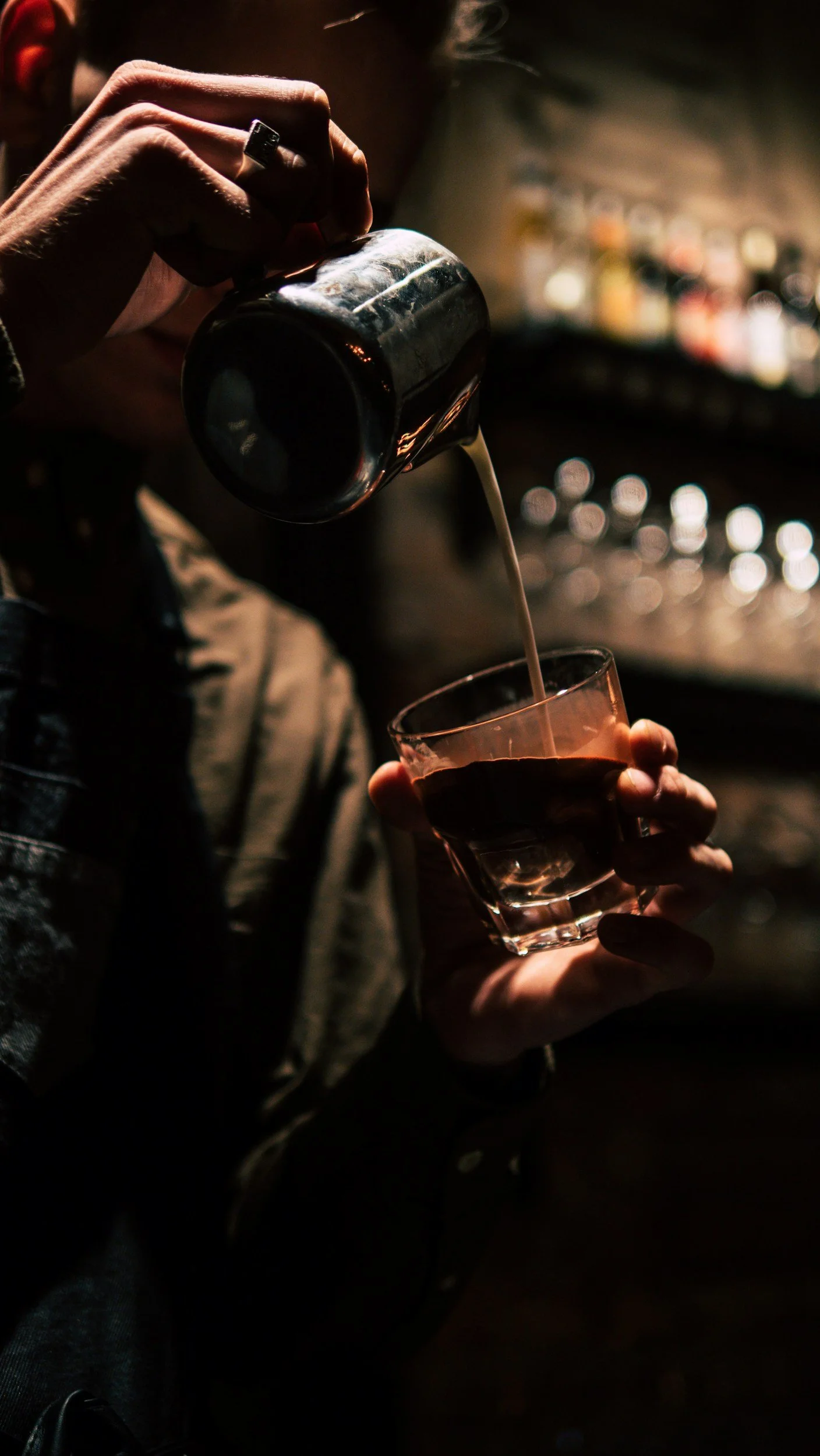 A person pouring a dark beverage, possibly coffee or espresso, from a container into a glass cup in a dimly lit bar or café setting.
