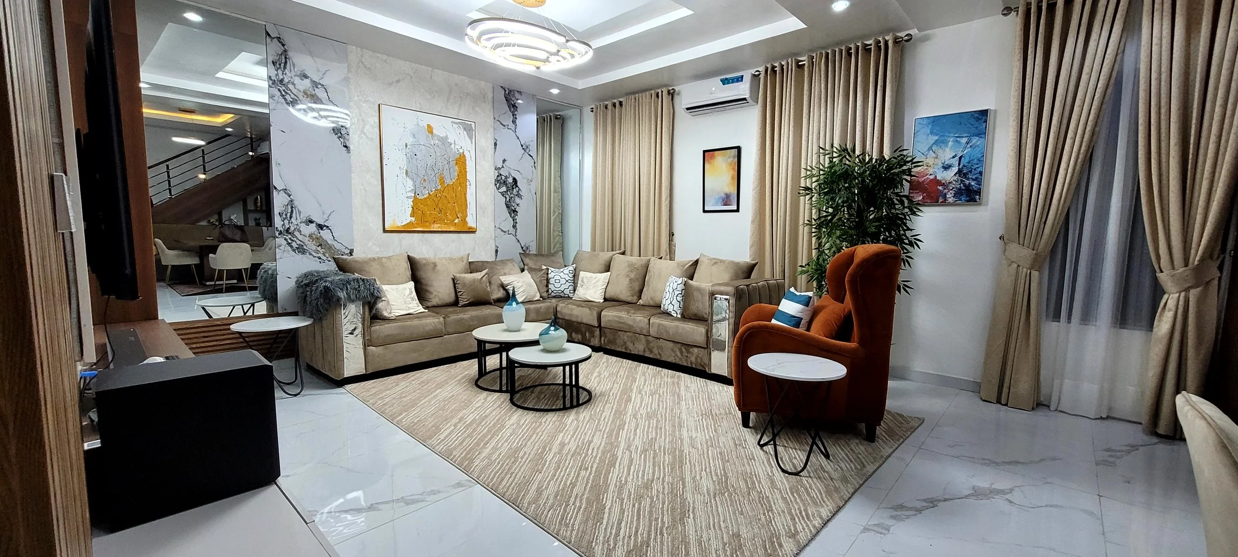 Modern living room with beige sectional sofa, orange armchair, and coffee tables with vases. Decor includes abstract art on wall, potted plant, curtains, and a light fixture on ceiling designed by IHOD.