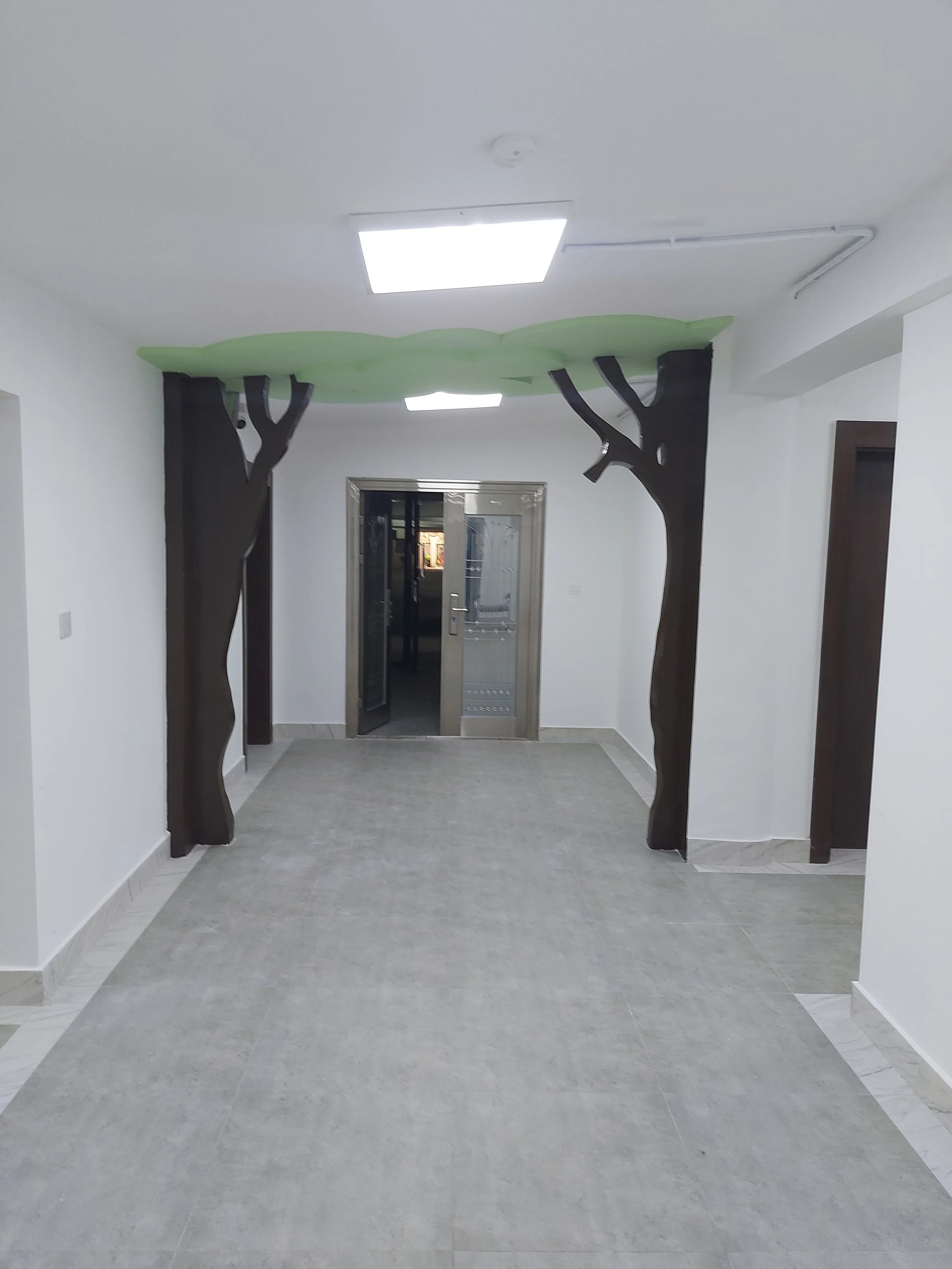 Hallway with decorative dark brown wooden tree-shaped supports on both sides, a reflective elevator door at the end, white walls, and a gray tiled floor, within the children’s church building (RCCG Olive Branch) Project Managed by IHOD.