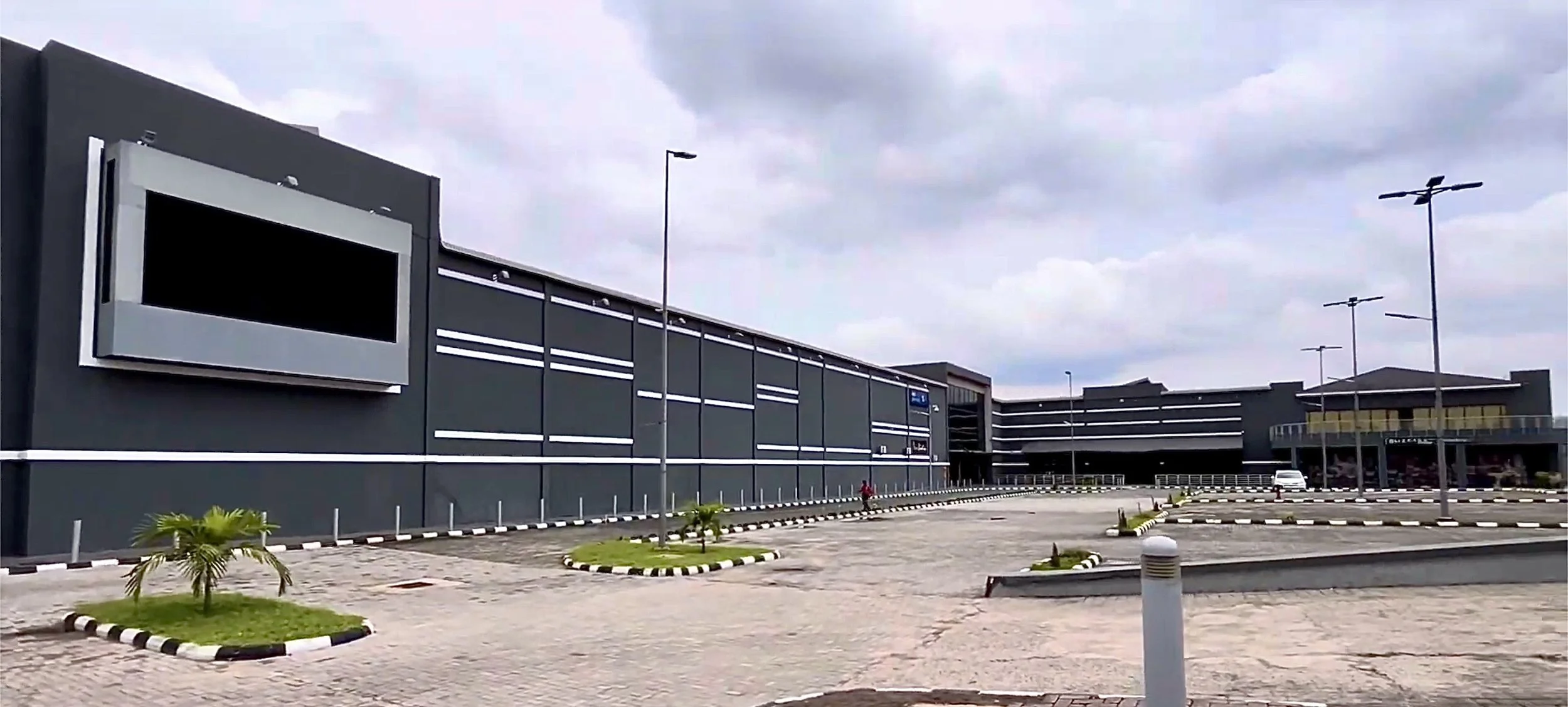 Modern shopping mall with parking lot, small landscaped areas with palm trees, street lights, and a few pedestrians under a cloudy sky.