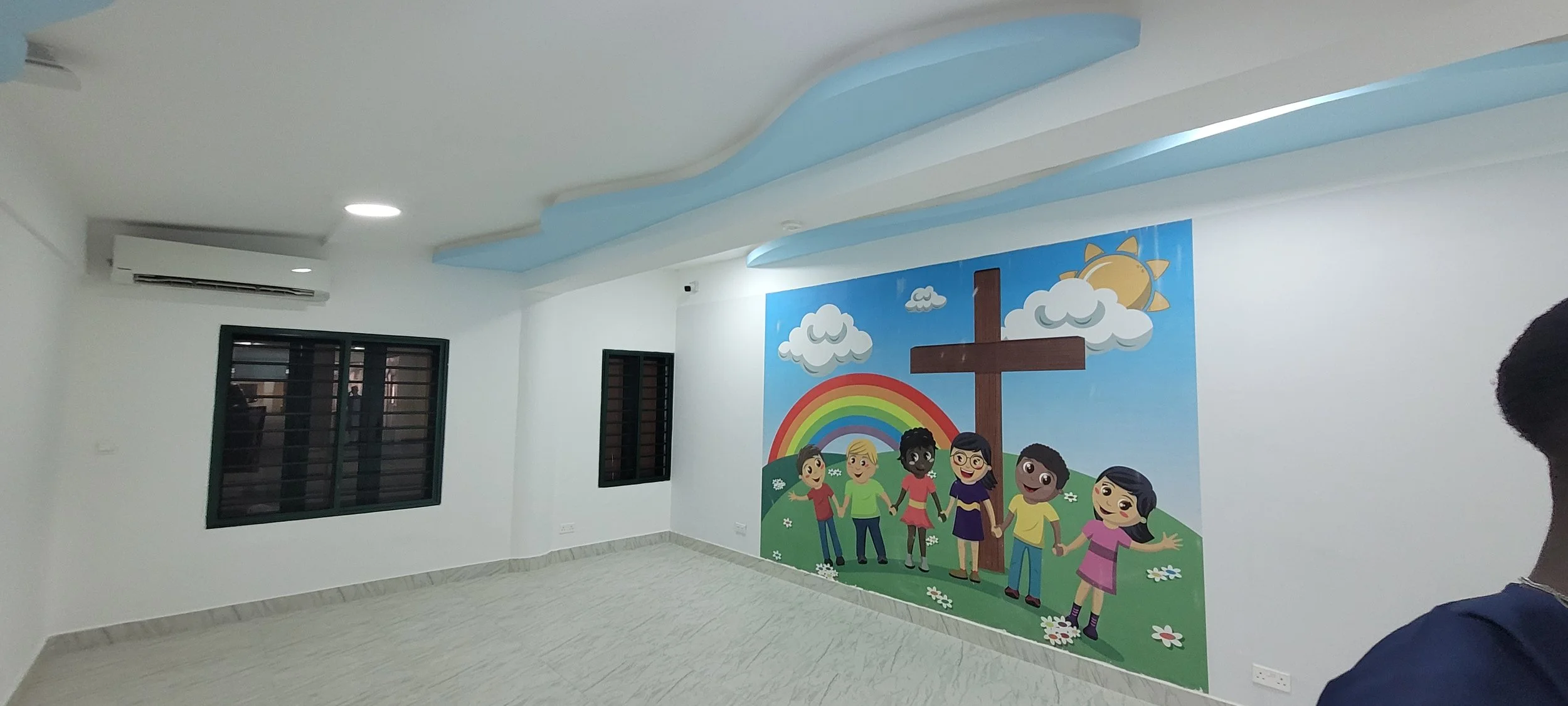 An indoor room with a large colorful mural of children holding hands around a wooden cross, surrounded by a rainbow, clouds, and a sun within the children’s church building (RCCG Olive Branch) Project Managed by IHOD.