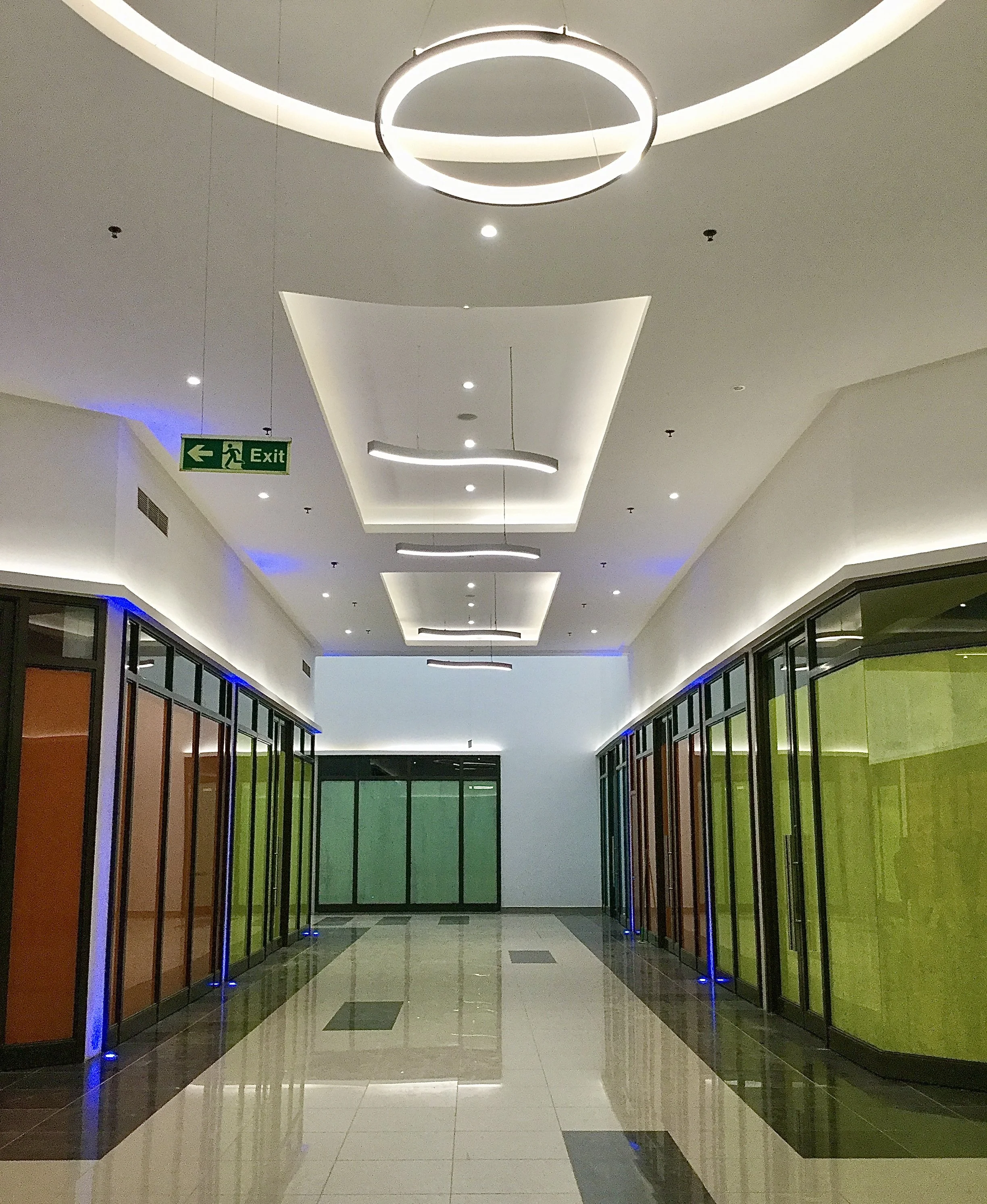 Interior of a modern shopping mall corridor with glossy tiled floors, colorful glass storefronts on both sides, and a ceiling with decorative lighting fixtures and an illuminated circular design. An exit sign with a running figure and an arrow pointing left is visible. The scene is well-lit and clean.