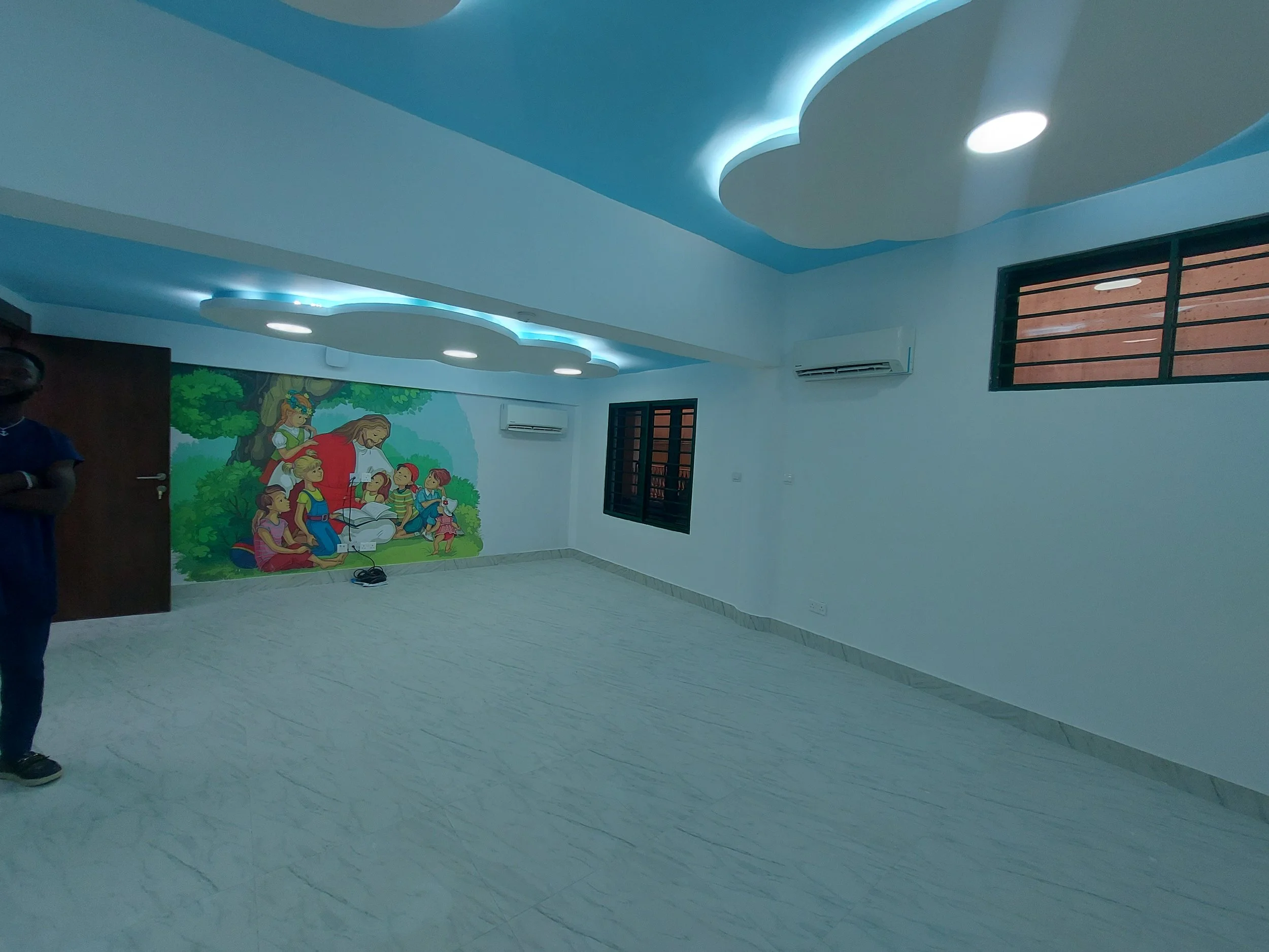 Empty room with blue ceiling, decorative ceiling lights, and a colorful mural of children and a woman reading under a tree on the wall, within the children’s church building (RCCG Olive Branch) Project Managed by IHOD.
