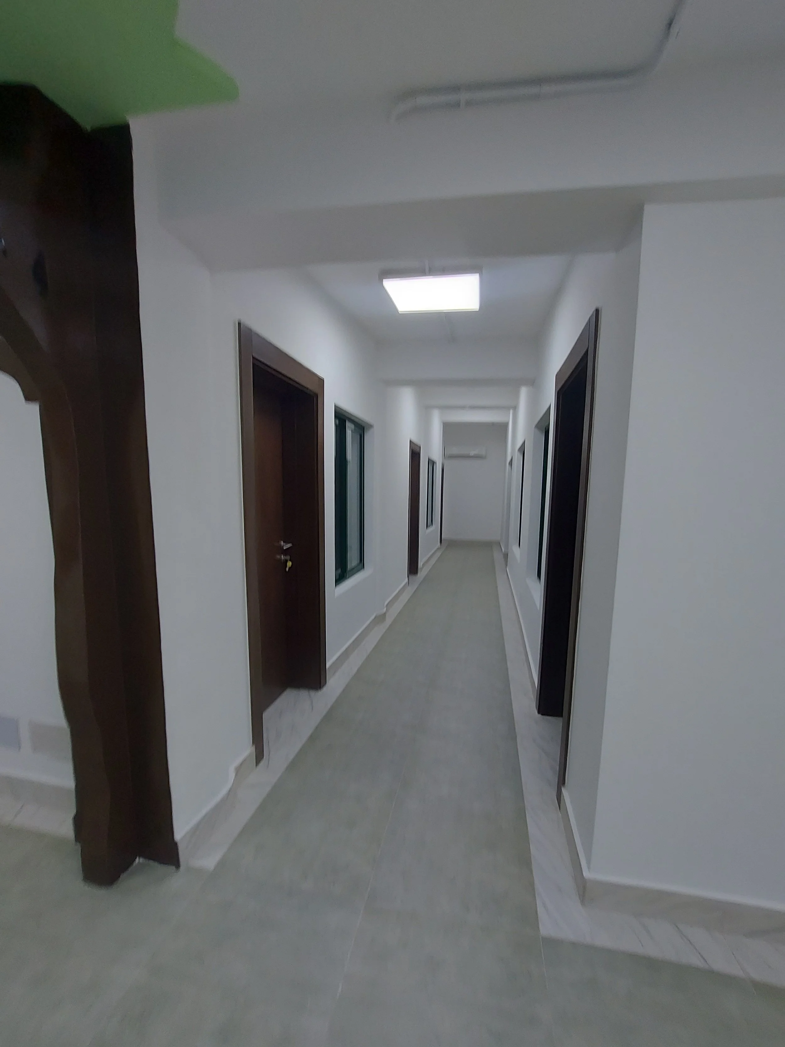 A long hallway with white walls, four doorways on the right side, two windows on the left wall, and a square ceiling light., within the children’s church building (RCCG Olive Branch) Project Managed by IHOD.