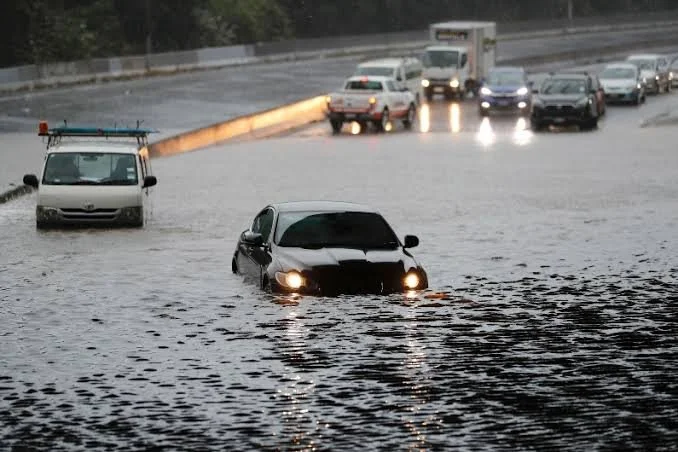 A black car driving through deep floodwaters on a flooded road during a heavy rainstorm, with other vehicles visible in the background.