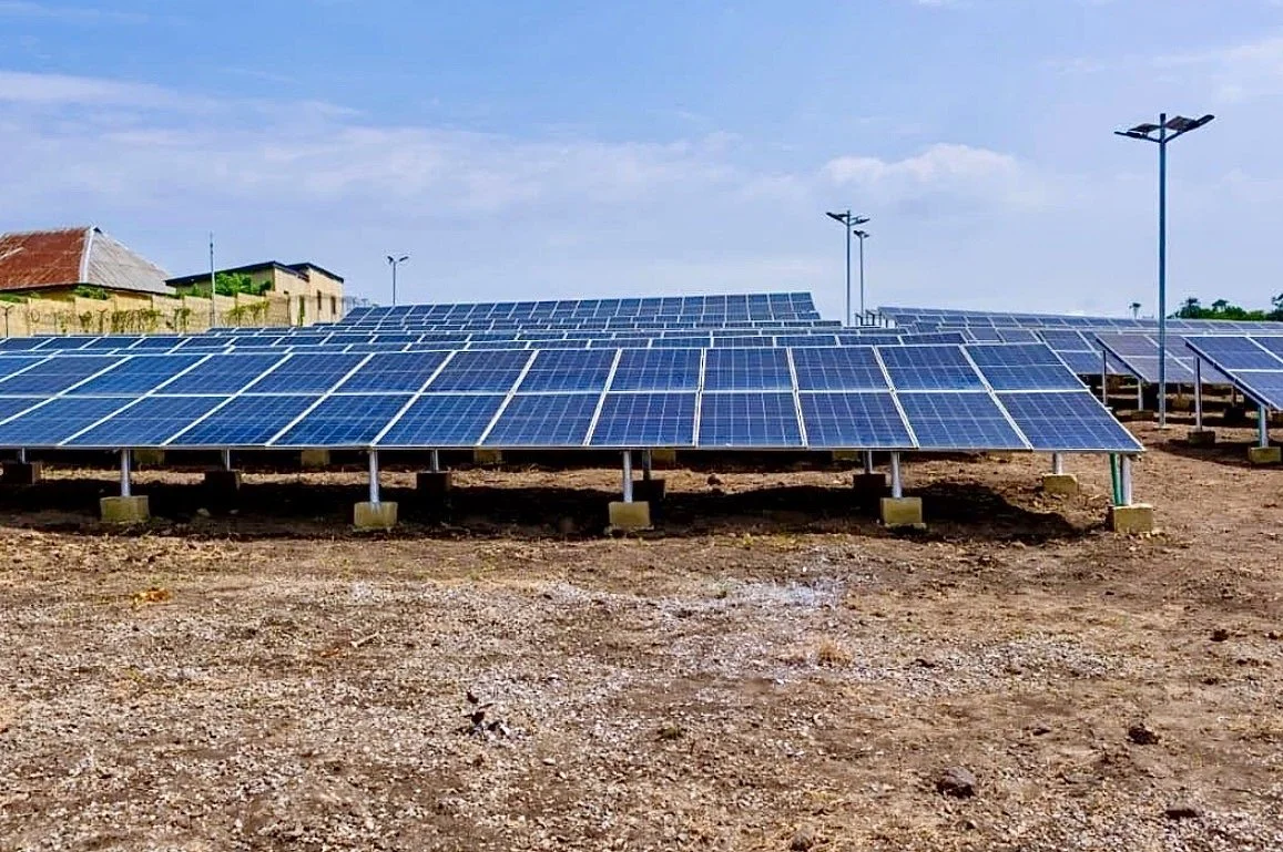 Solar panels installed on a dirt ground in an open area, with small buildings and lamp posts in the background under a blue sky.
