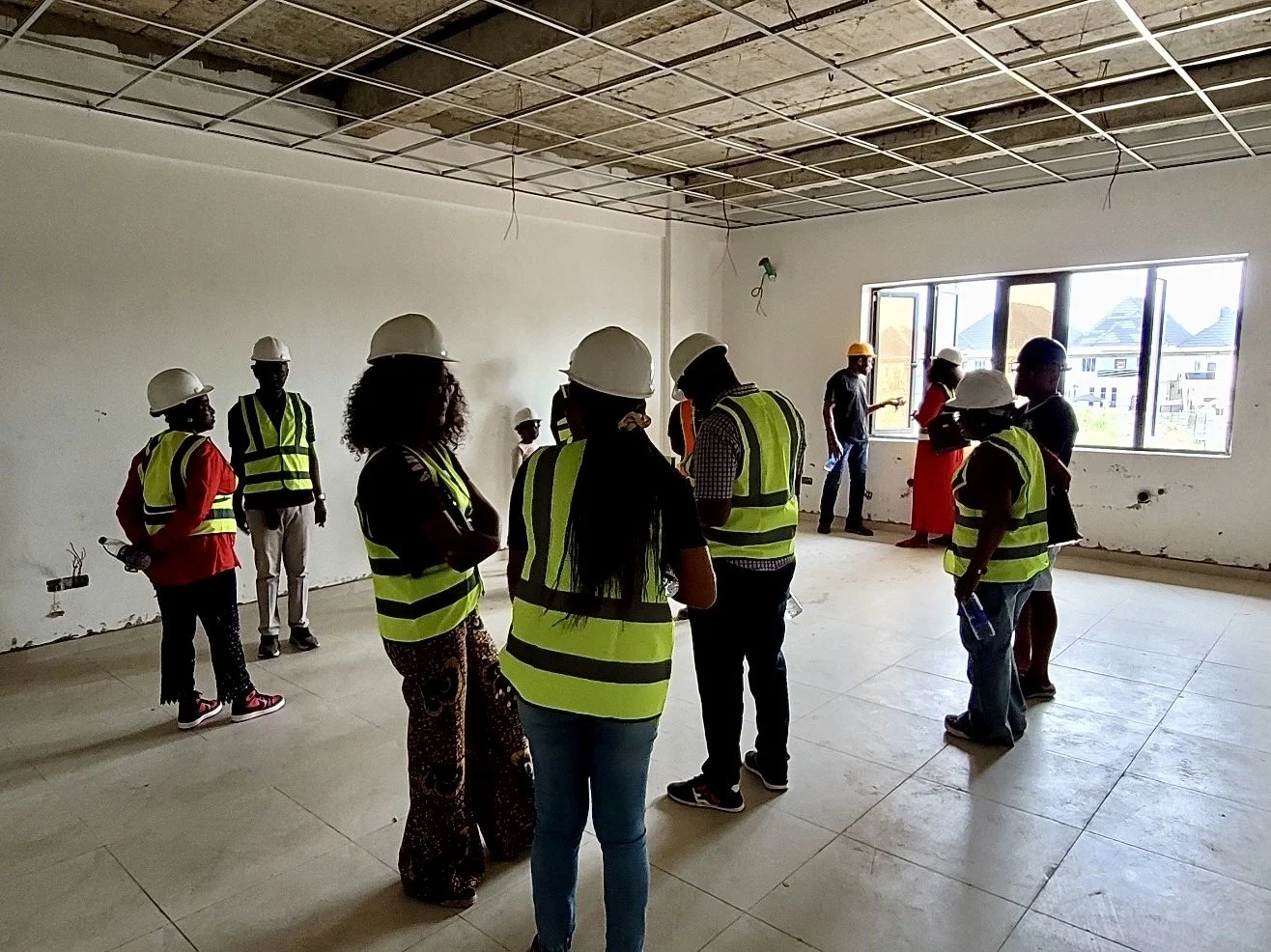 Group of construction workers and supervisors inside a building under construction, wearing safety vests and helmets, with some looking at the surroundings and others talking.