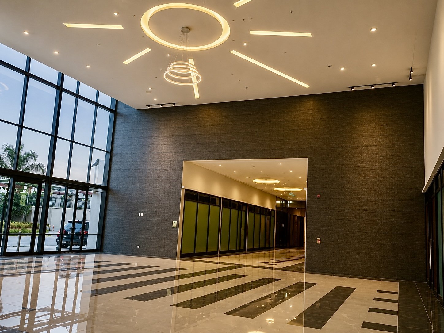 Modern hotel lobby with large glass windows, high ceiling, decorative lighting, dark textured wall, and checkered marble floor.