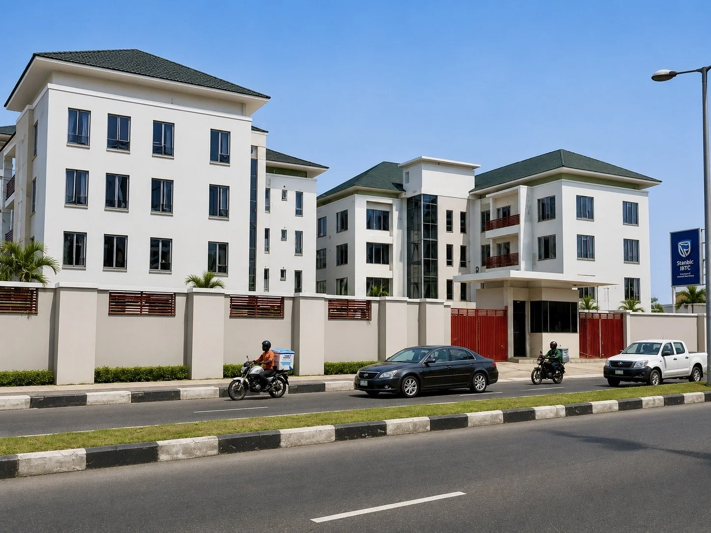 Modern white apartment complex with multiple floors, green roofs, and glass windows, surrounded by a high wall. In front, a street with cars and motorcycles, and a blue sign indicating Stanbic IBTC bank. Clear blue sky overhead.