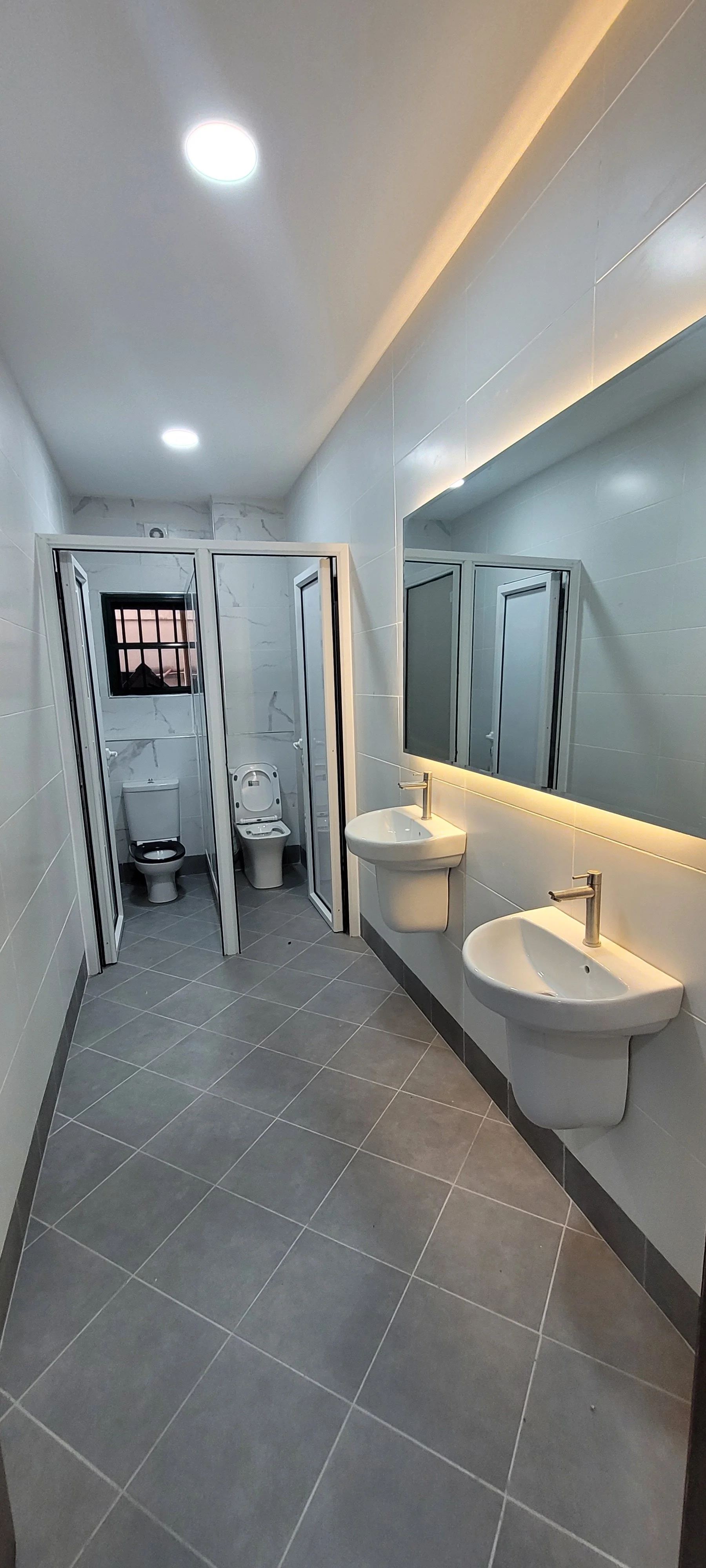 A modern commercial bathroom with two sinks and a large mirror, two toilets behind a glass partition, gray floor tiles, and white walls with an illuminated mirror, within the children’s church building (RCCG Olive Branch) Project Managed by IHOD.
