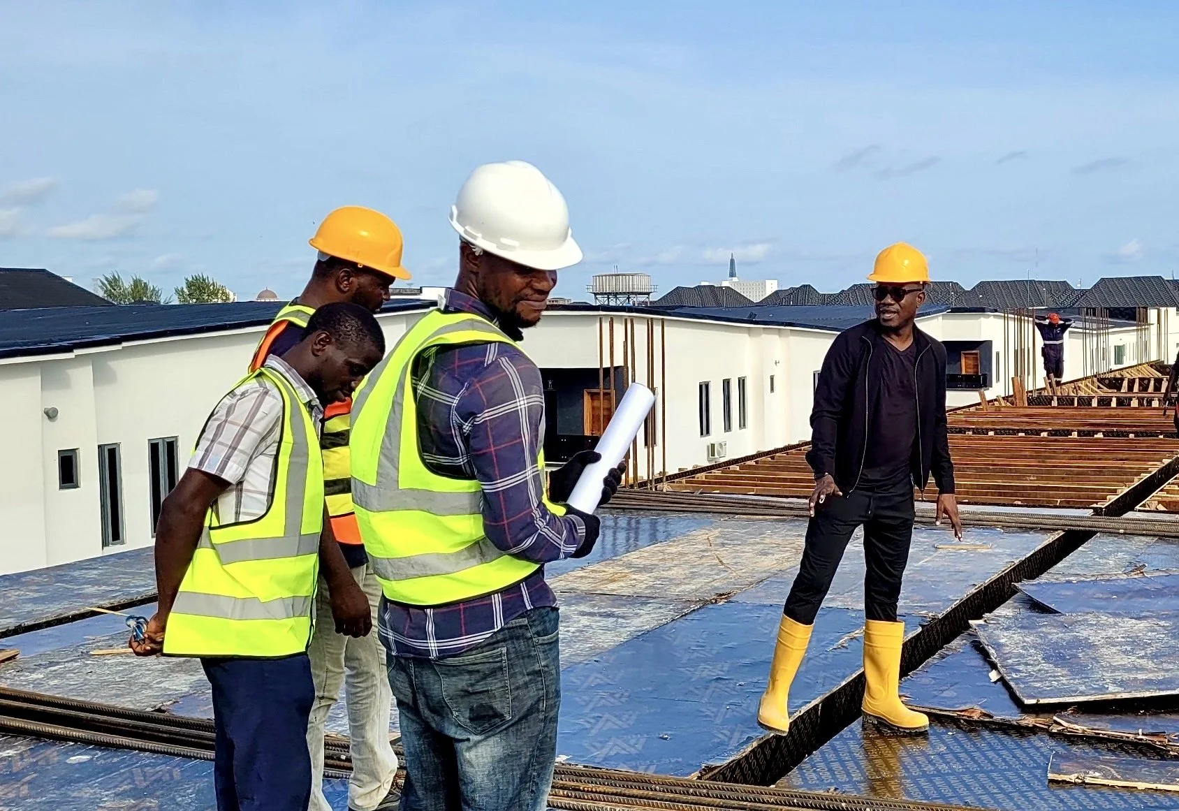 Construction workers inspecting a building site, some wearing safety vests and helmets, one holding a blueprint, with a partially built structure and town in the background.