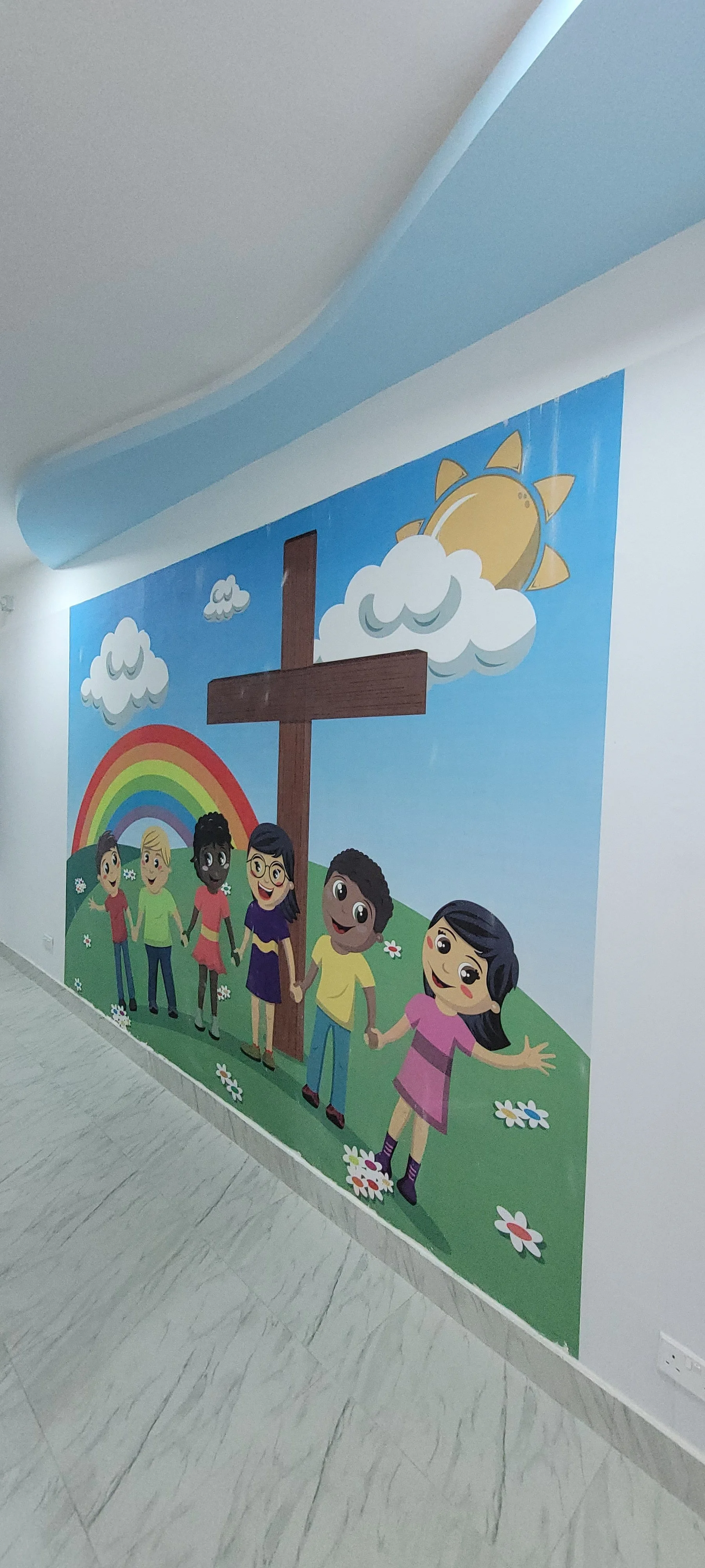 Colorful mural of children holding hands around a cross, with rainbow, smiling sun, and clouds in a bright sky, outside on a white wall, within the children’s church building (RCCG Olive Branch) Project Managed by IHOD.