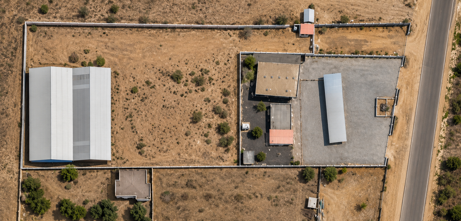 An aerial view of a fenced property with multiple buildings, gravel and paved areas, and surrounding dry grass land. The property is bordered by a road on the right side.
