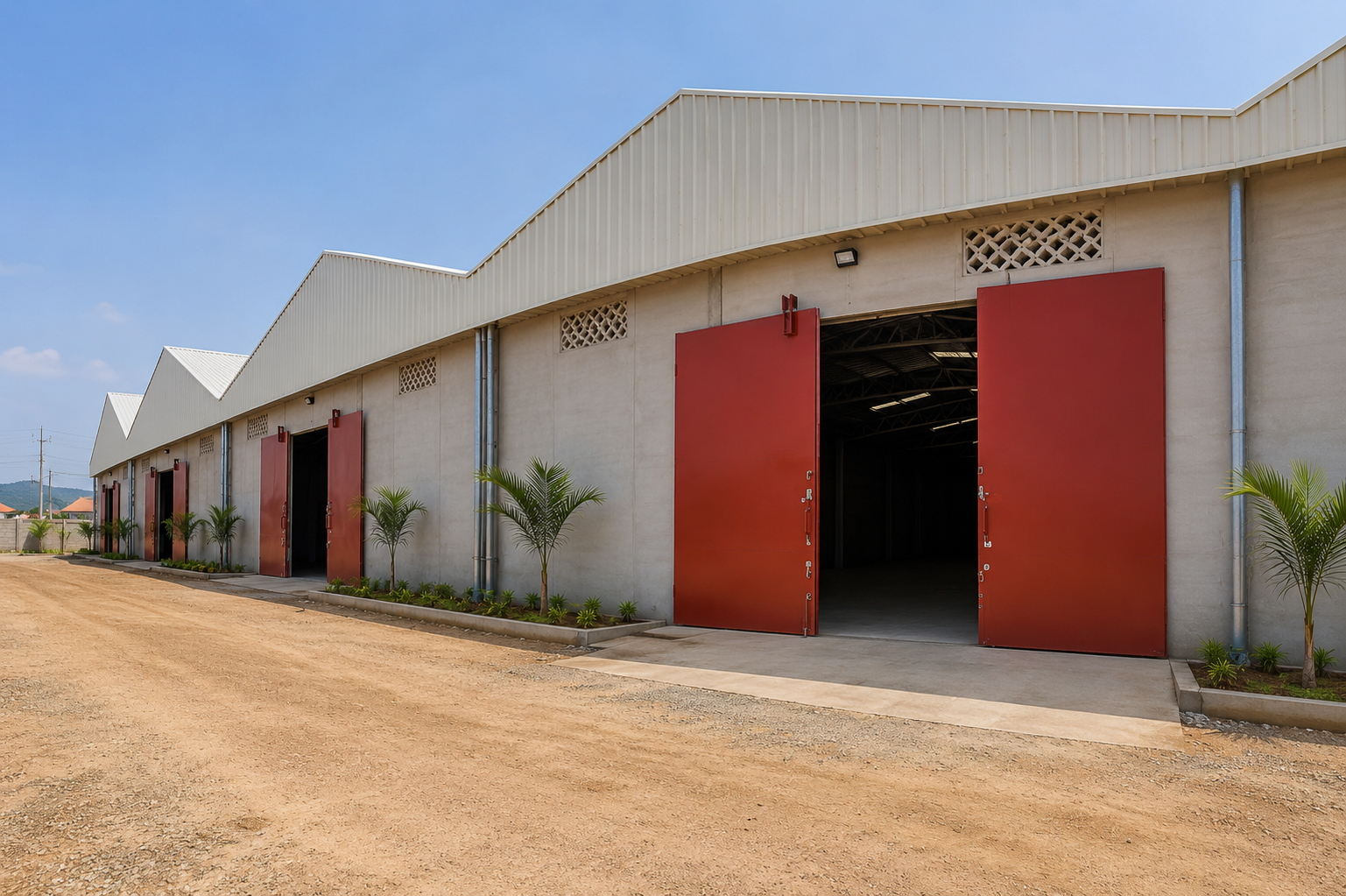 Industrial warehouse with open red doors, small palm trees lining the exterior, and a dirt road in front, under a blue sky.