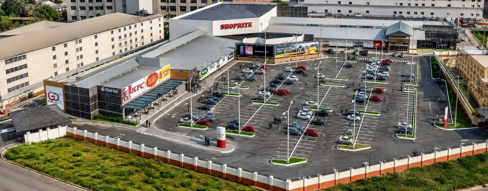 A large shopping mall with a big parking lot, featuring several cars parked and some people walking. The store signs include KFC, Pizza Inn, and ShopRite. The mall has multiple entrances, and there are streetlights throughout the parking lot.