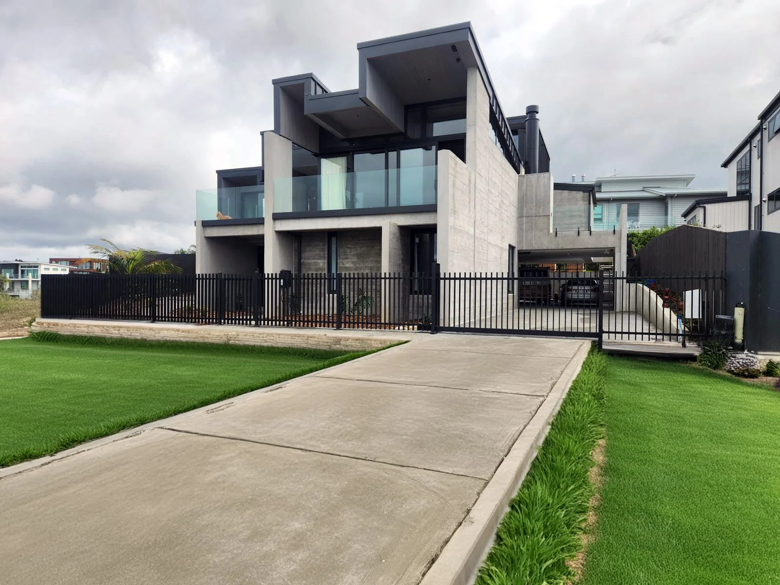 Modern multi-story house with concrete and glass features, black metal fence, driveway with parked cars, and well-manicured green lawn.