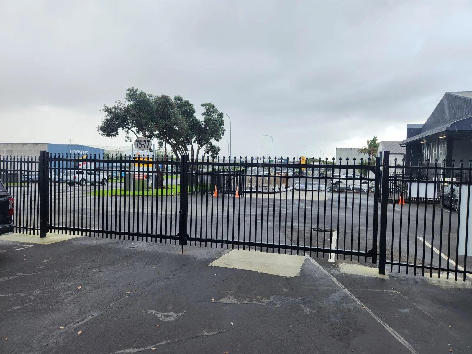 A black metal gate with a parking lot and commercial buildings in the background on a cloudy day.