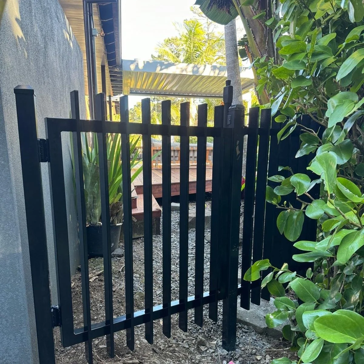 Black metal gate in front of a garden area with plants and a deck. Part of a house with an overhang and trees in background.