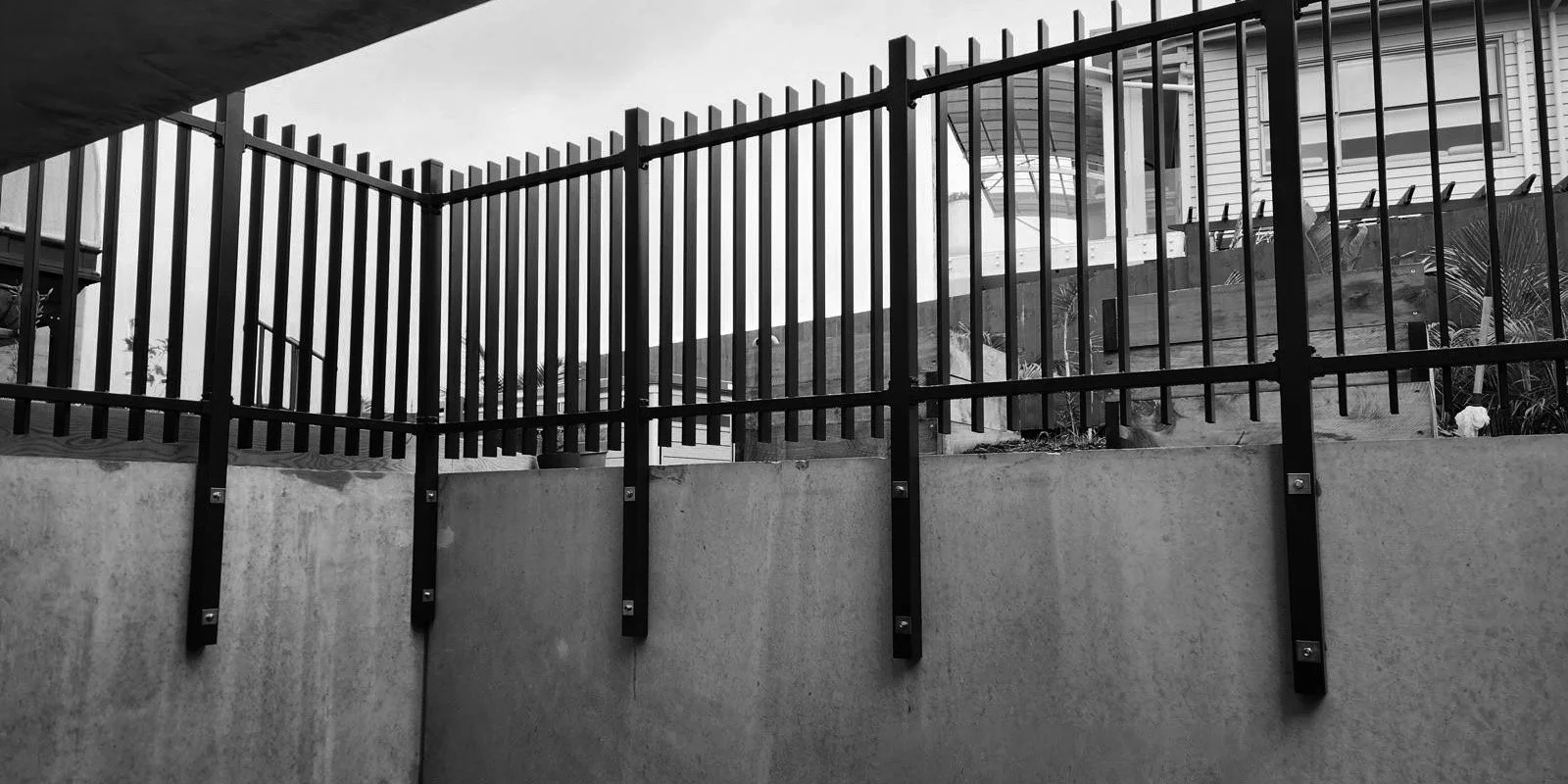 Black metal fence on a concrete wall in front of a house with stairs and a deck, black-and-white photo.