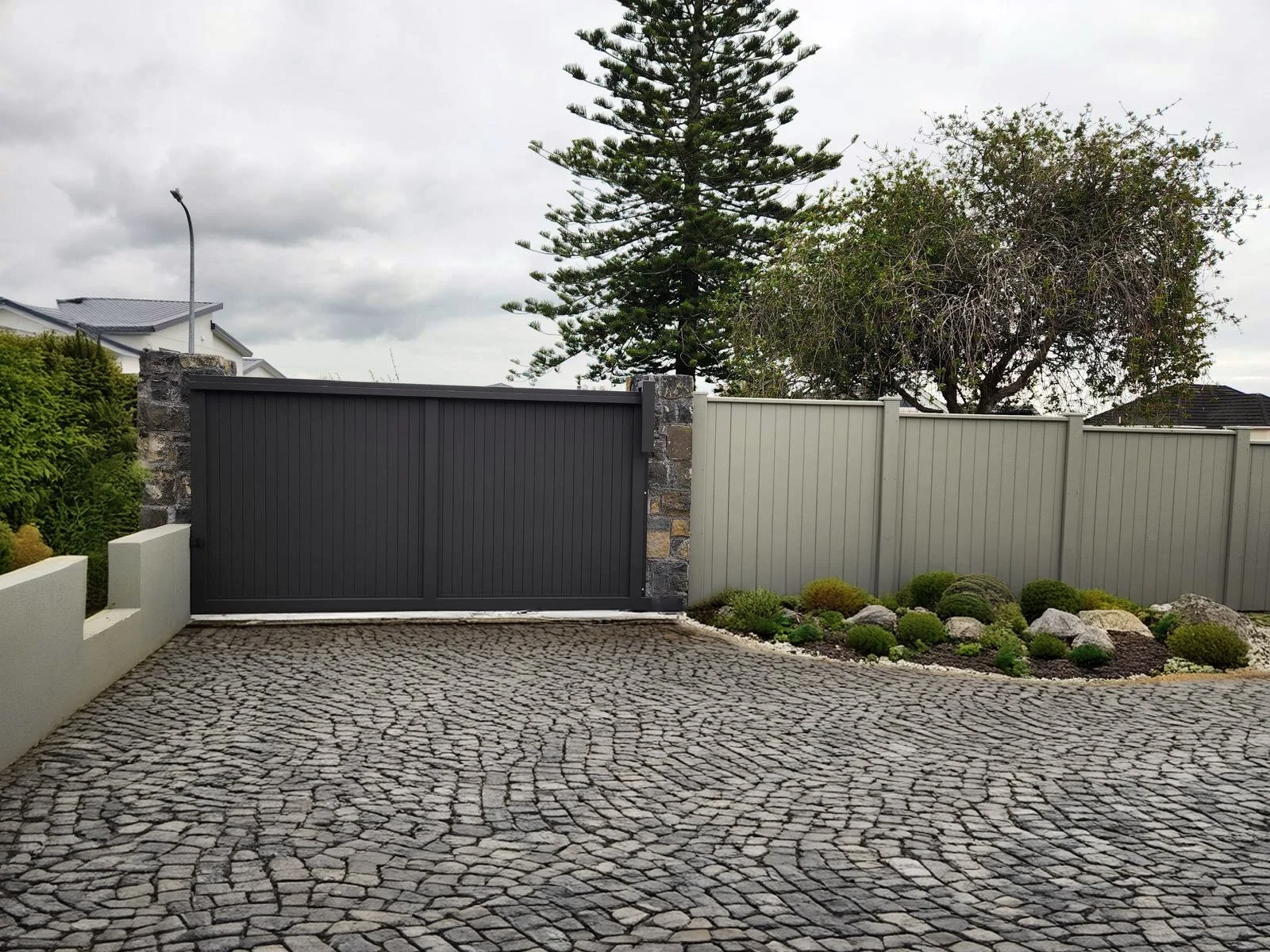 A driveway with cobblestone paving, bordered by a low white concrete wall and a garden bed with rocks and small bushes on the right. Behind the driveway is a grey and white metal fence with stone pillars, with tall trees and cloudy sky in the background.