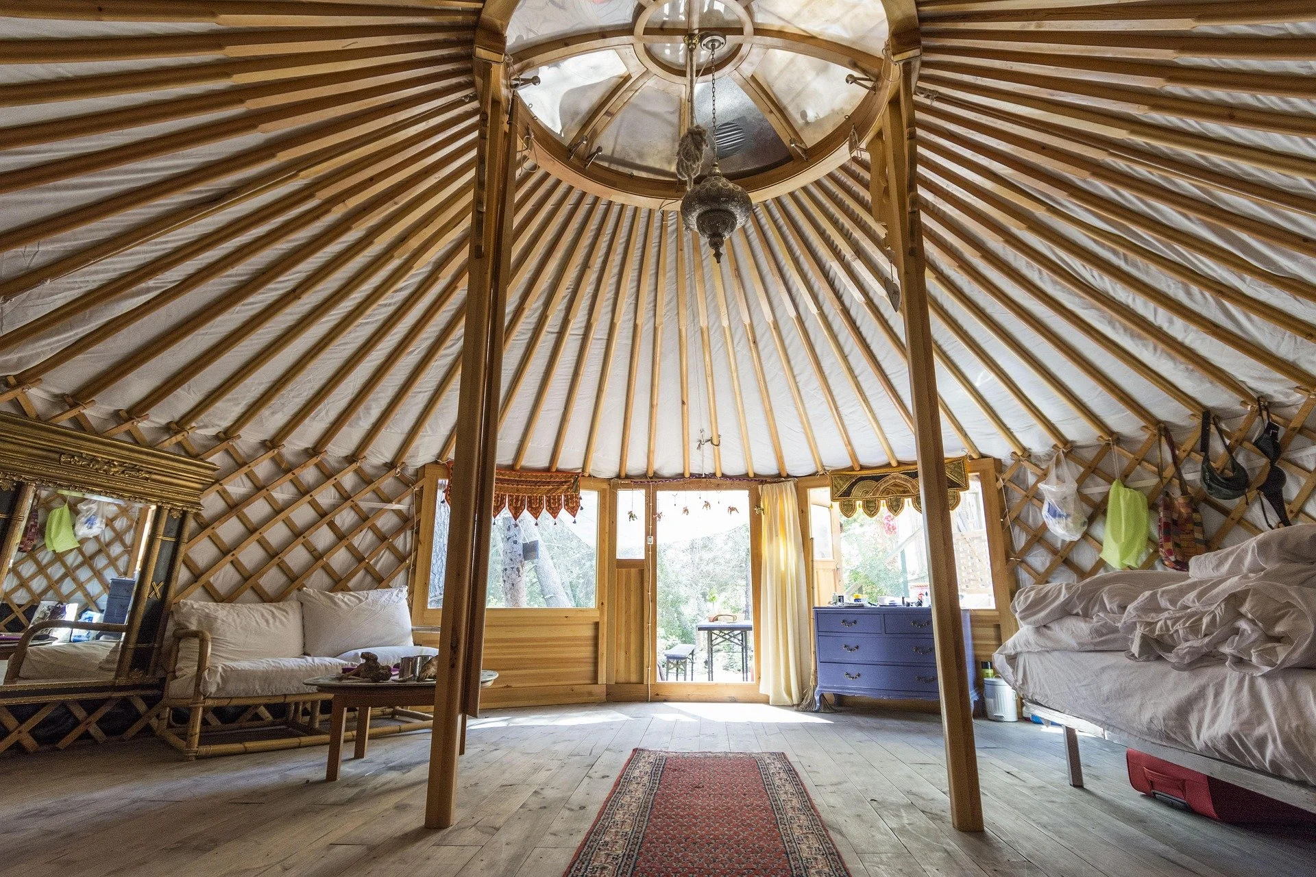 Interior of a yurt with a wooden lattice structure, a central chandelier, windows letting in natural light, and furniture including a sofa and a bed.