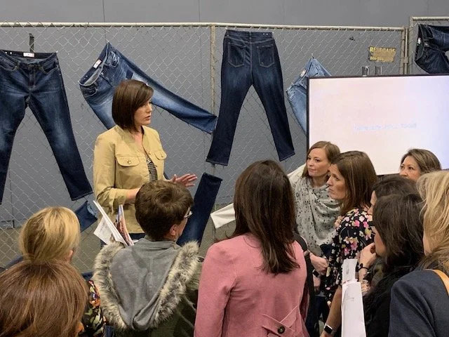 Julie Ann Beijer in a tan jacket talks to a group of women in a room with denim jeans hanging on a chain-link fence behind her and a whiteboard to her right.