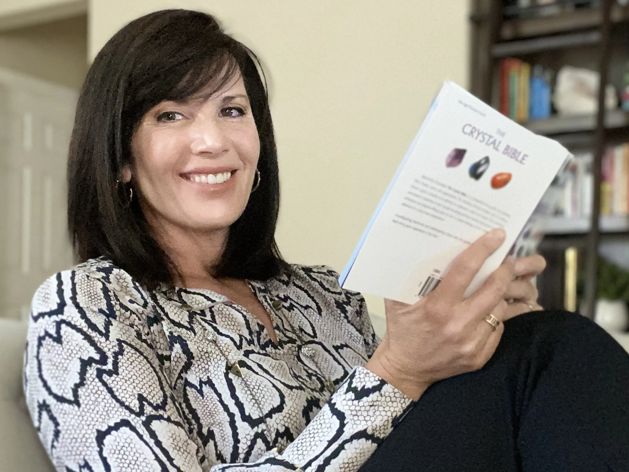 Julie Ann Beijer holding a book titled 'The Crystal Bible' in a room with shelves of books in the background.