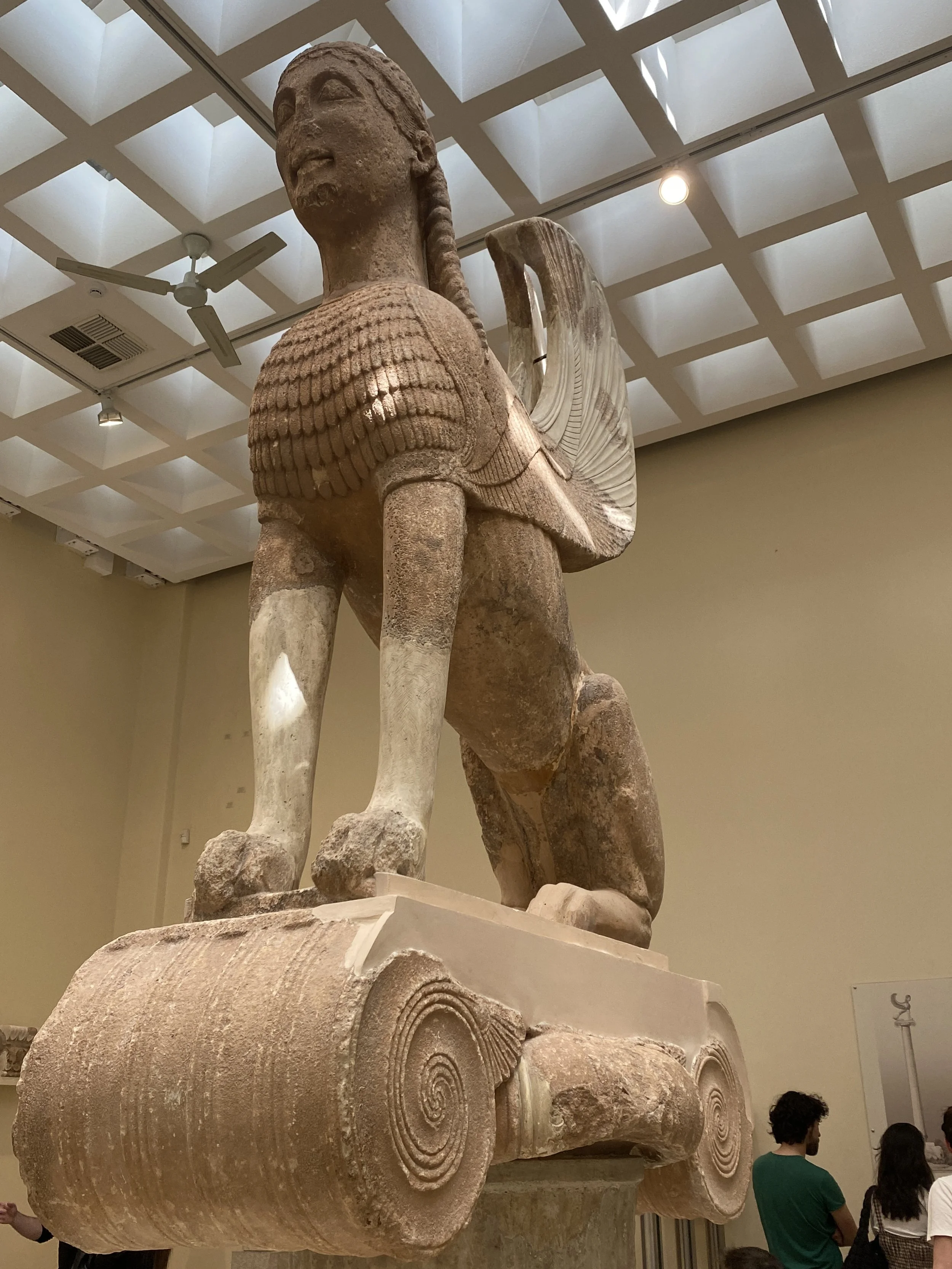 Ancient stone sculpture of a mythological creature with the body of a lion and the head of a human, displayed in a museum, with visitors observing it.