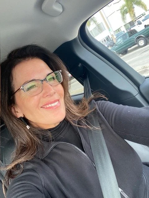 Woman with glasses smiling in the driver's seat of a car, holding the camera for a selfie, with a street and parked cars visible through the window.