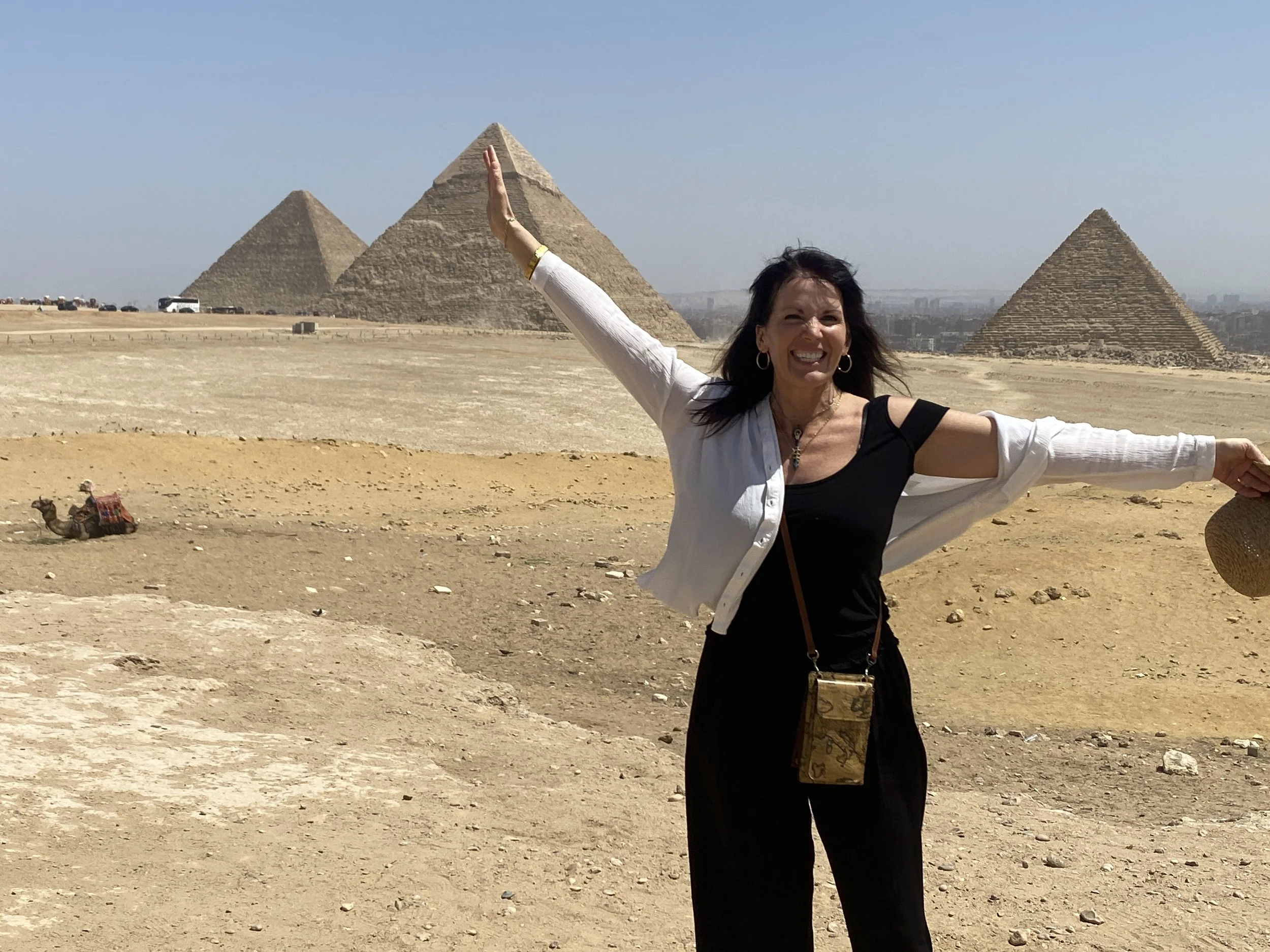 Julie Ann Beijer with her arms outstretched, standing in front of the pyramids of Egypt under a clear blue sky. She is wearing earrings, a necklace, and a white jacket over black clothing, with a small purse hanging around her neck.