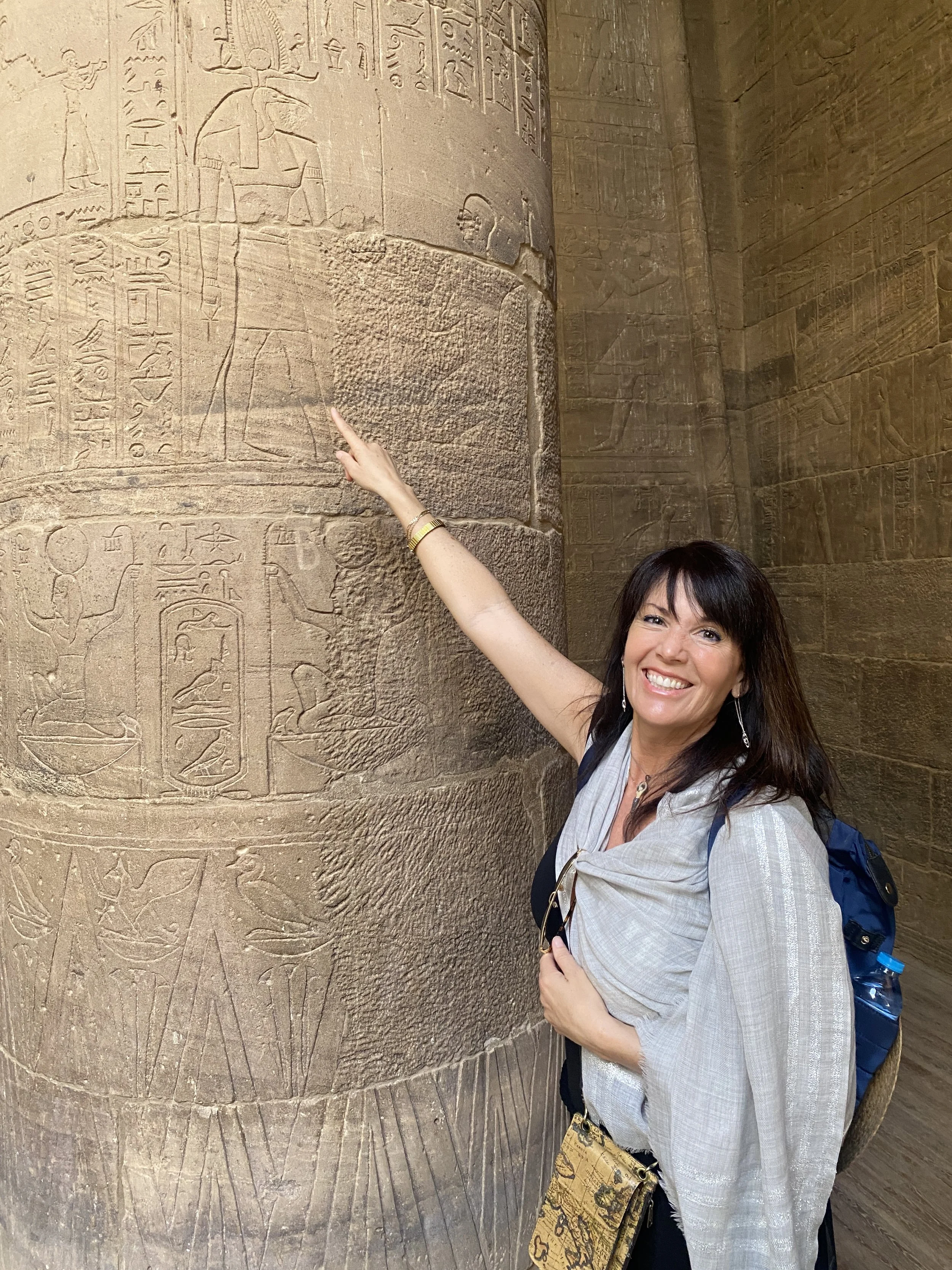 Julie Ann Beijer smiling and pointing at ancient Egyptian hieroglyphs and carvings on a stone wall at Karnak, Egypt. She has dark hair, is wearing a light-colored shirt, and has a shoulder bag on.