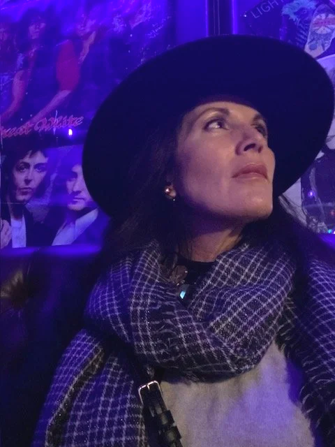 Julie Ann Beijer wearing a large black hat and a checkered scarf, sitting indoors with purple lighting and posters in the background.
