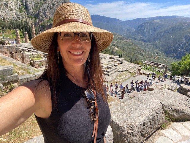 Julie Ann Beijer taking a selfie at Delphi, in Greece, an outdoor archaeological site with ruins and a mountain landscape in the background.