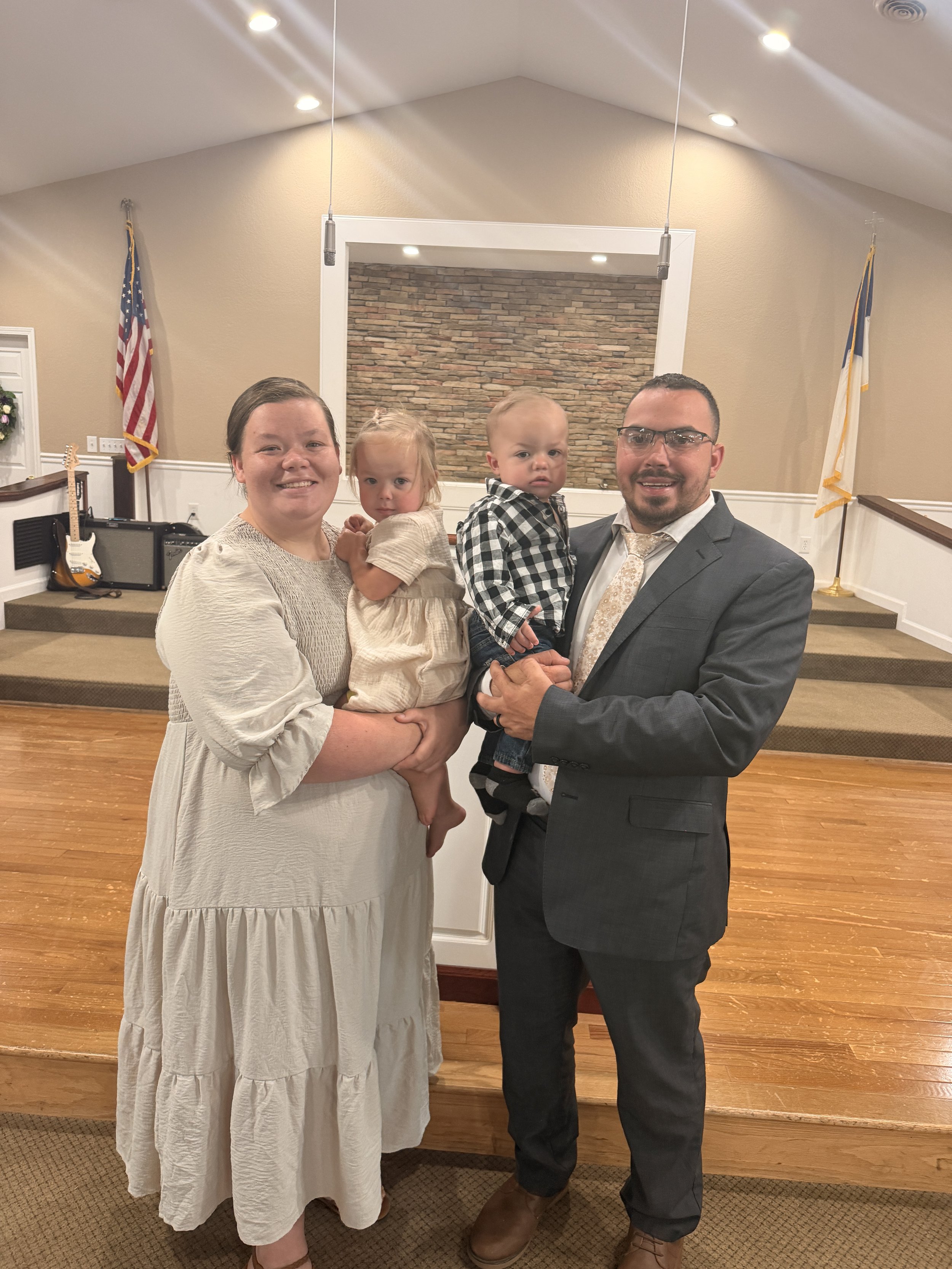 A smiling woman in a white dress holds a young girl in a beige dress, while a man in a gray suit and glasses holds a young boy with a checkered shirt, inside a room with a brick wall backdrop, American flags, and guitars on the side.