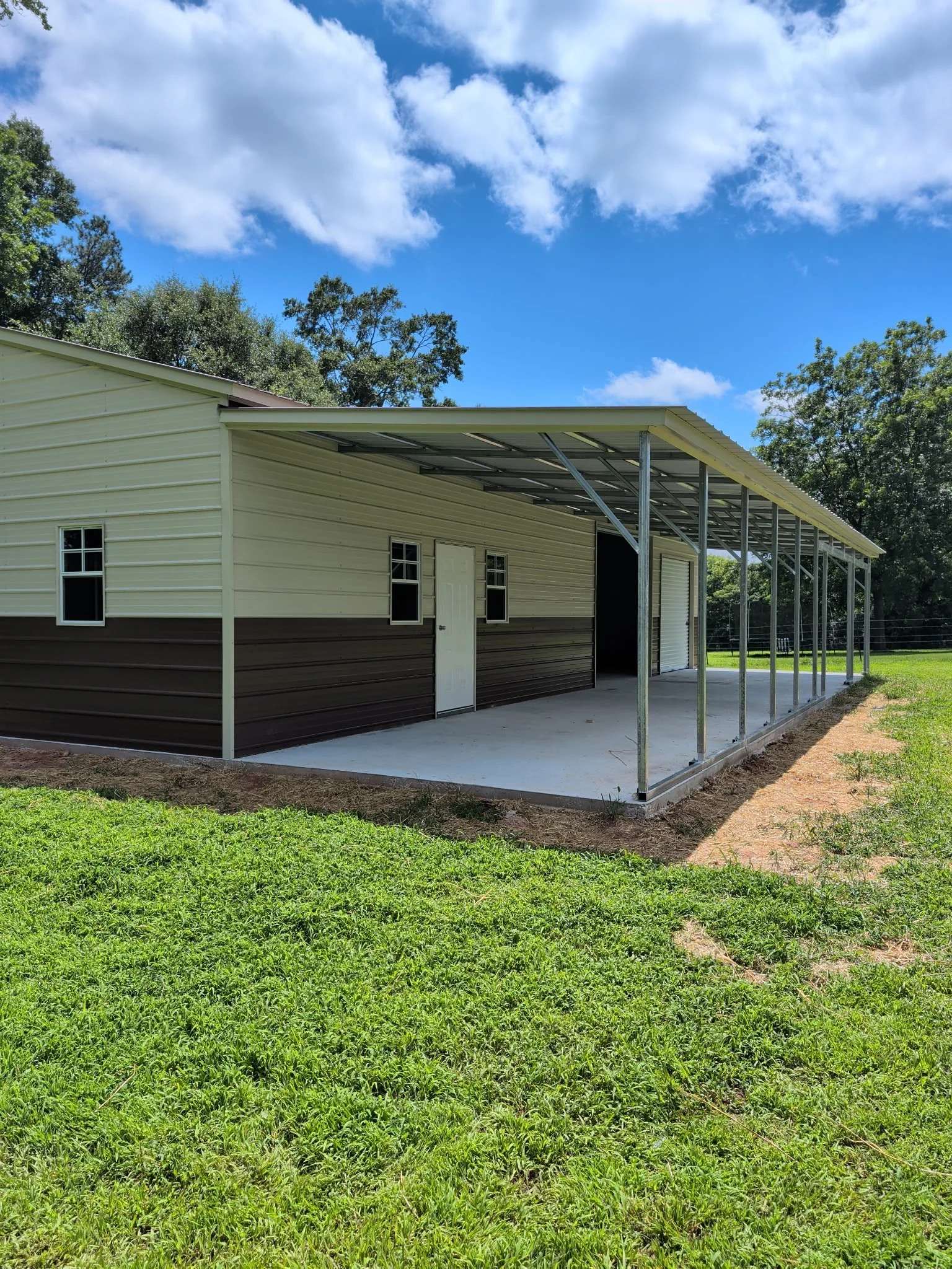 A partly constructed metal building with a roof and partial sidewalls, set on a grassy area with trees and a blue sky with clouds in the background.